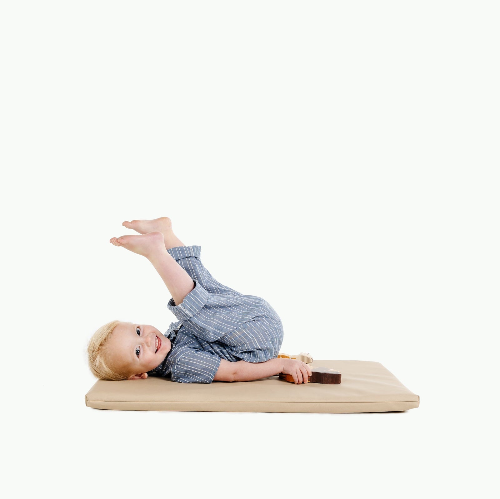 A young child lies on a Gathre Wholesale Padded Changing Mat in Millet, smiling and playfully raising their legs, holding a rectangular object. The mat rests on a plain white background.