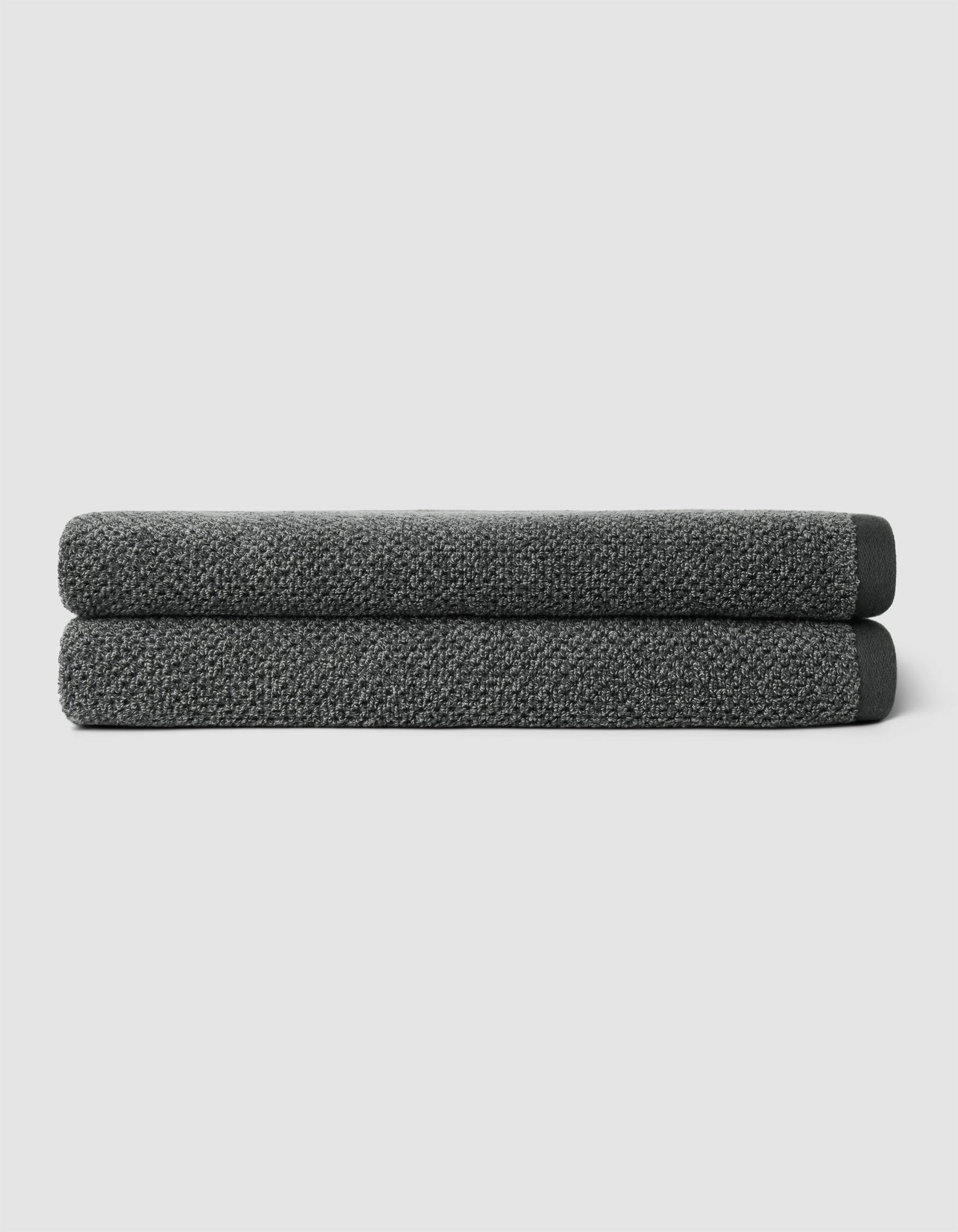 Two neatly folded Nantucket Bath Towels by Cozy Earth, in dark gray, are stacked atop one another against a plain light gray background.|Color:Graphite