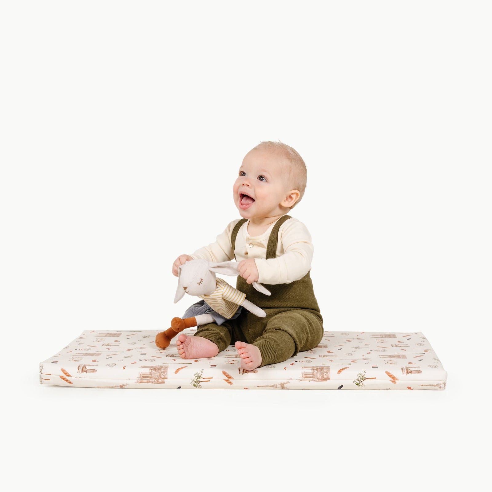 A smiling baby in green overalls sits on the Gathre Paris Wholesale Padded Changing Mat, holding a stuffed animal toy, against a white background.