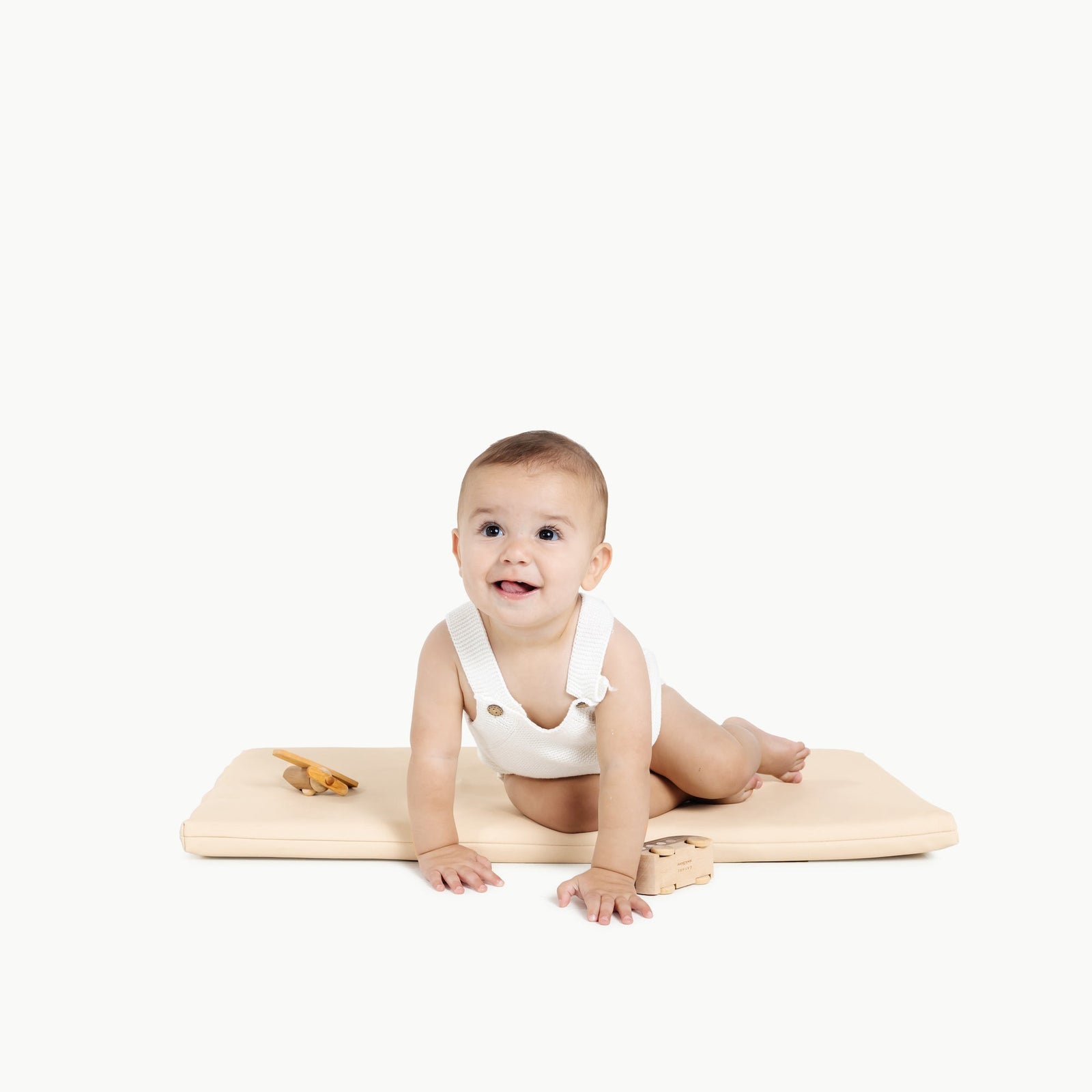 A smiling baby in a white onesie crawls on the Gathre Wholesale Padded Changing Mat in Petal, with wooden toys nearby, against a plain white background.