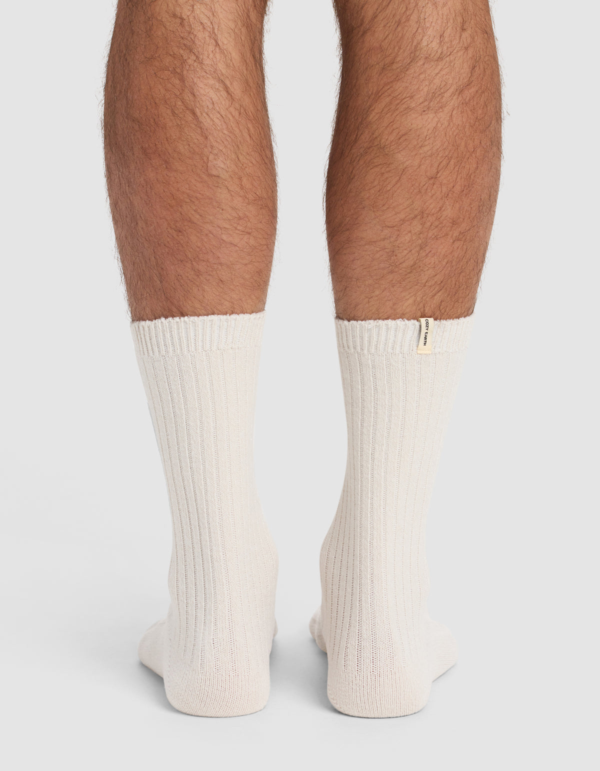 Back view of a person's lower legs wearing The Plush Lounge Sock 3-Pack by Cozy Earth—thick, ribbed, off-white socks with a small beige tag near the top edge. Plain, light-colored background.