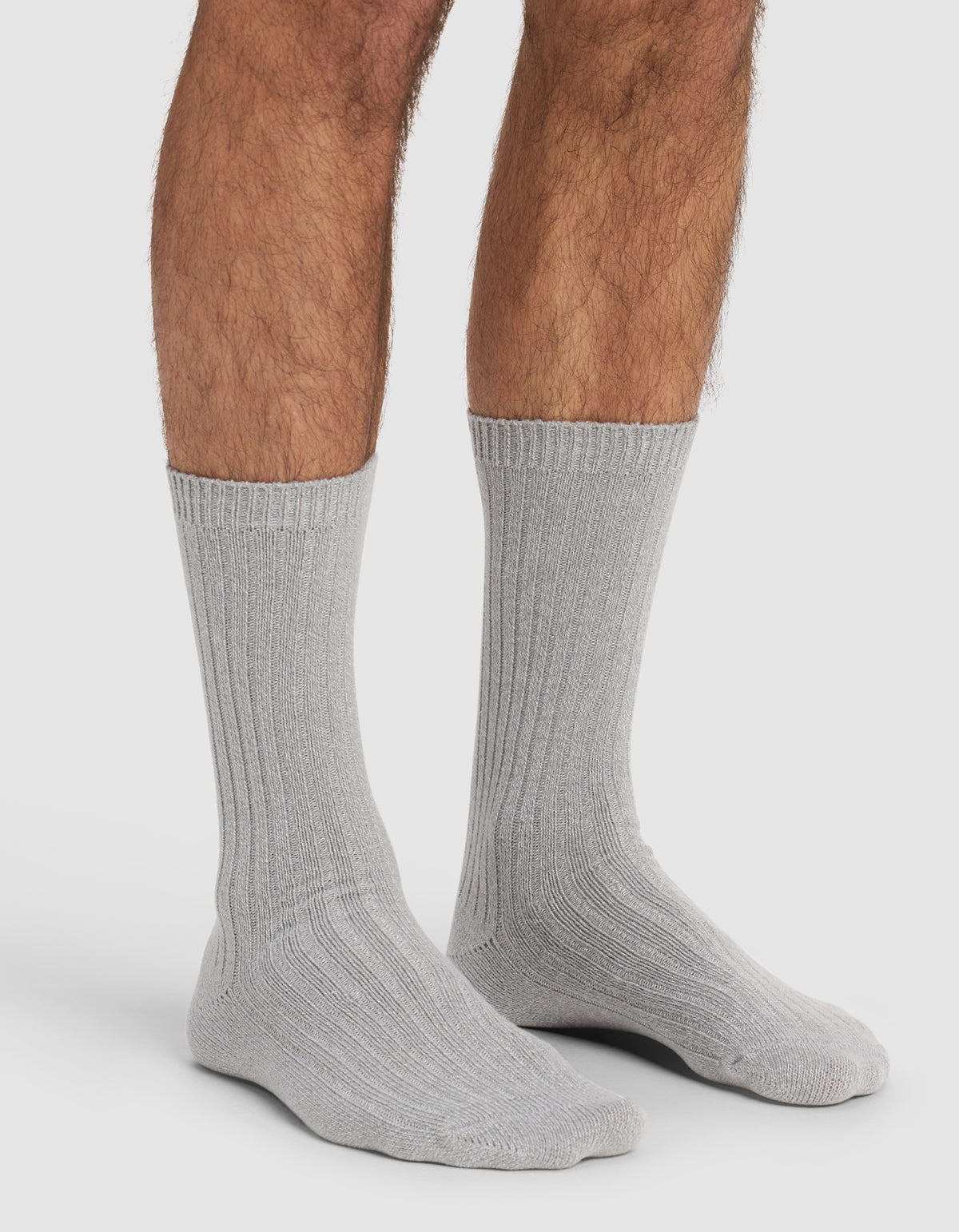 A person wears Cozy Earth's The Plush Lounge Sock 3-Pack in light gray, ribbed crew style, with only their lower legs and feet visible against a plain white background.