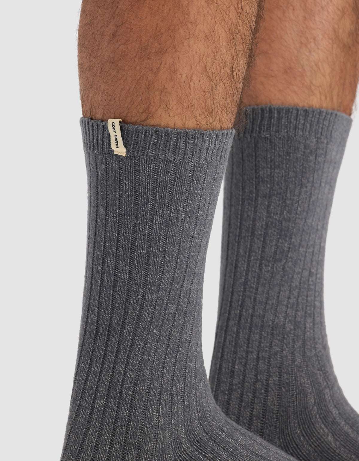 Close-up of a person's lower legs wearing The Plush Lounge Sock in dark gray from Cozy Earth, featuring ribbed texture and a small beige tag near the top. Plain white background.