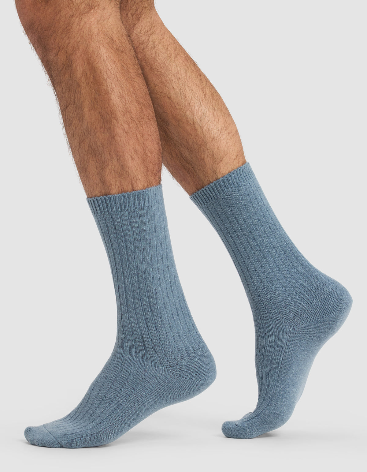 A person with hairy legs models Cozy Earth's The Plush Lounge Sock 3-Pack in light blue, ribbed crew style, standing on a plain white background with one foot flat and the other slightly raised, toes pointed.