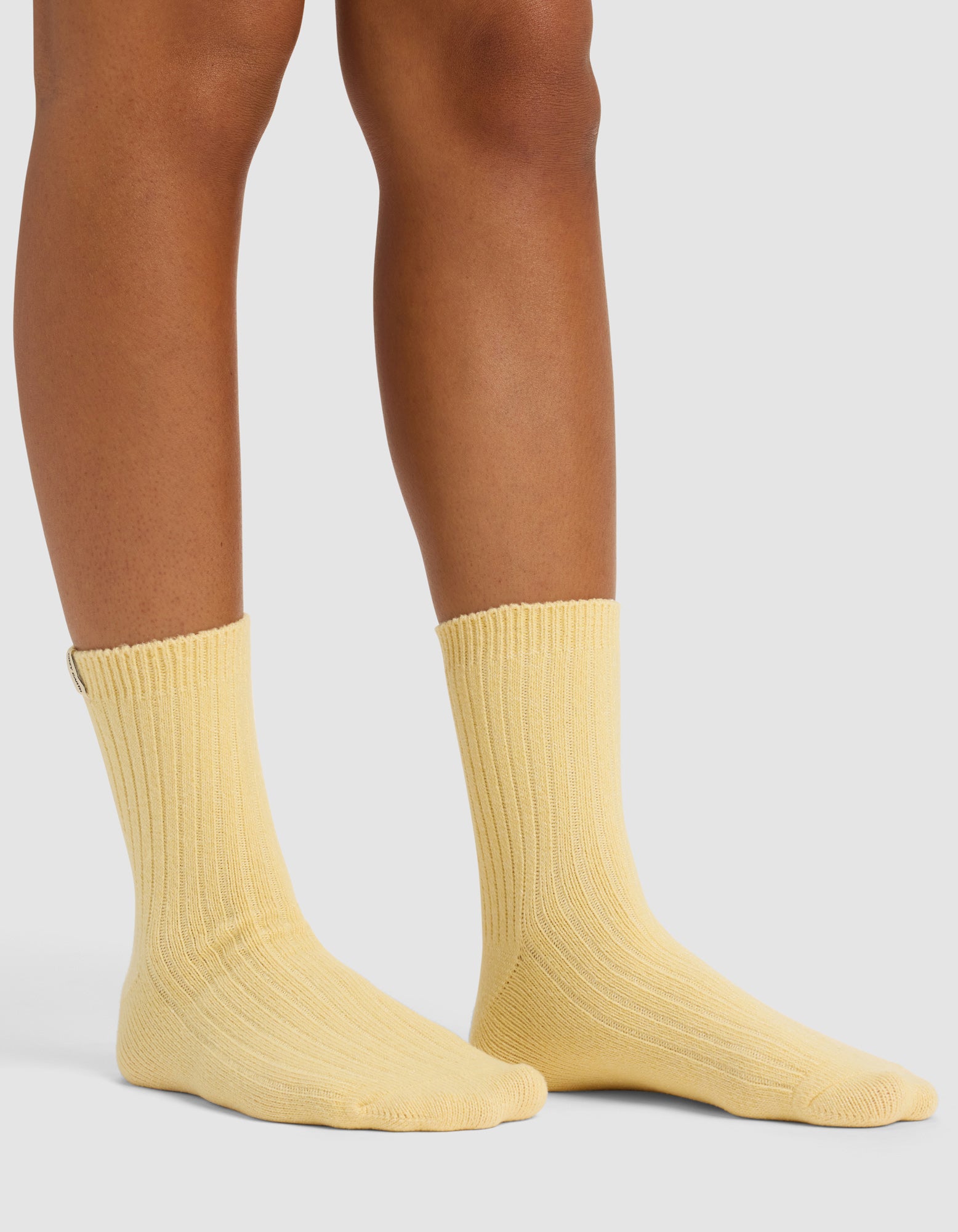 Someone wearing The Plush Modern Crew Sock by Cozy Earth in light yellow stands on a pale surface, with only their lower legs and feet visible.|Color:Daisy