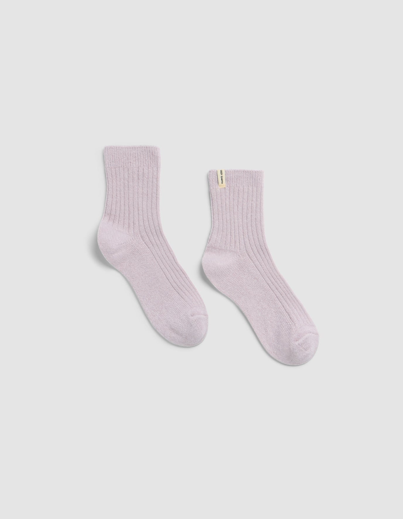 The Plush Modern Crew Sock by Cozy Earth in light pink features a ribbed design and a small yellow tag on one sock, shown against a plain white background.