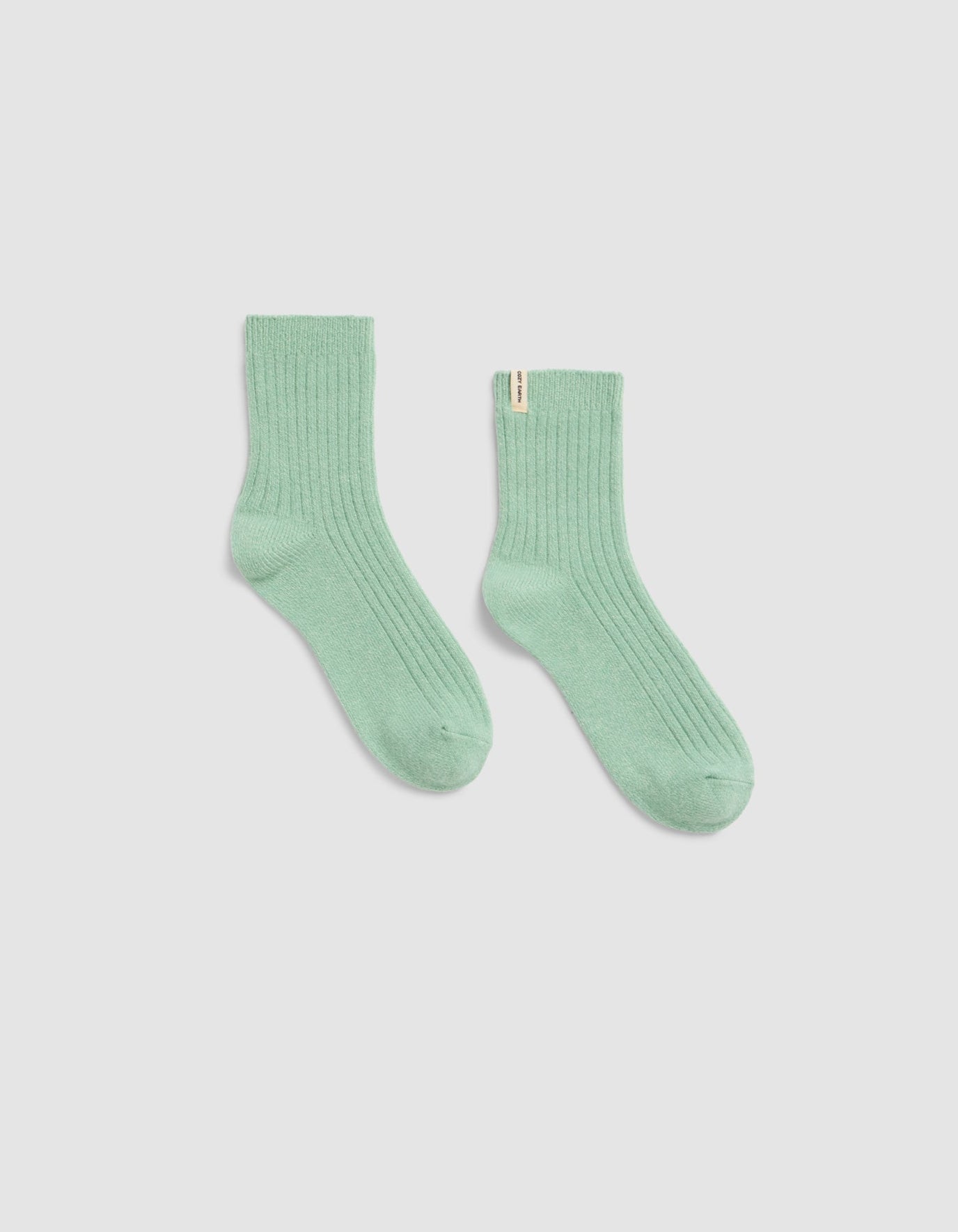A pair of The Plush Modern Crew Socks by Cozy Earth, in light green ribbed fabric, is laid flat on a plain white background with one sock slightly angled outward.