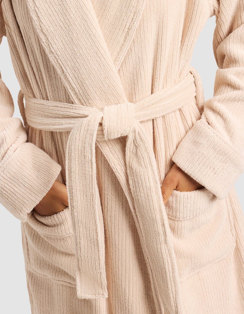 A person wears the Cozy Earth Ribbed Terry Bath Robe in beige, featuring a textured finish, waist belt, and hands in the front pockets.