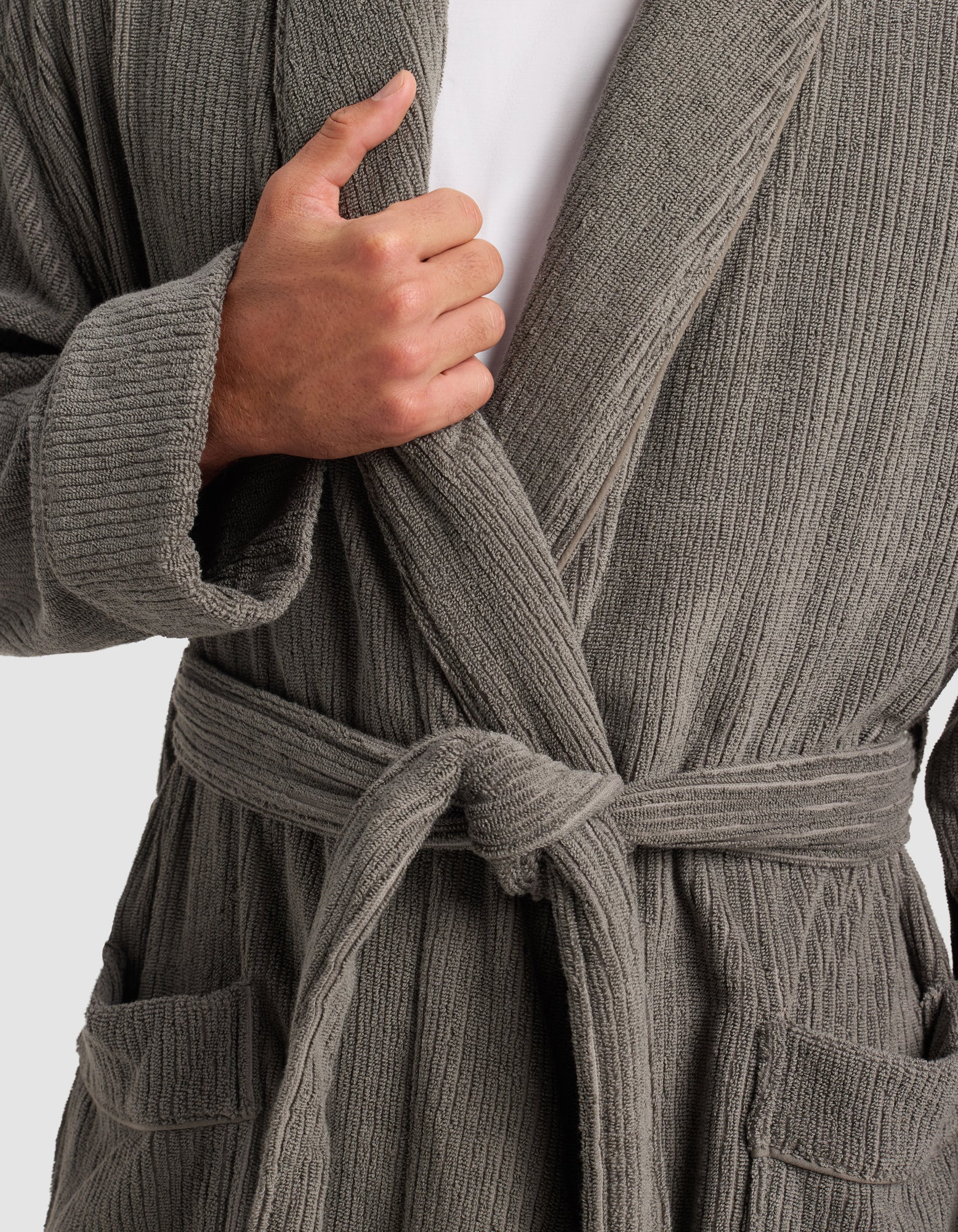 A person models Cozy Earth's Ribbed Terry Bath Robe in gray, tied at the waist, with one hand holding the lapel. The upper body, shirt underneath, and robe pockets are visible.