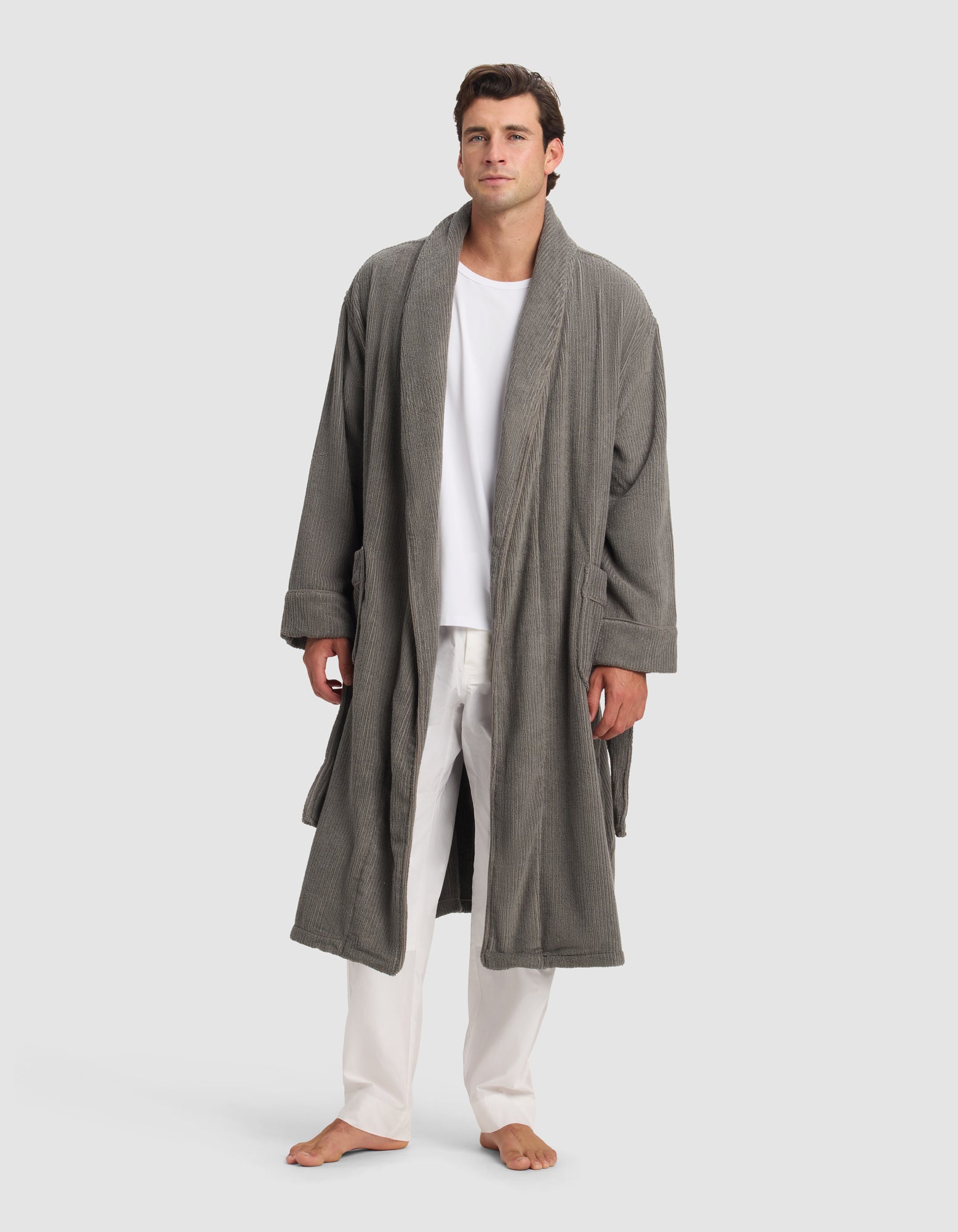 A man stands barefoot against a plain light background, wearing Cozy Earth’s Ribbed Terry Bath Robe over a white shirt and white pants. His hands rest at his sides as he looks straight ahead.