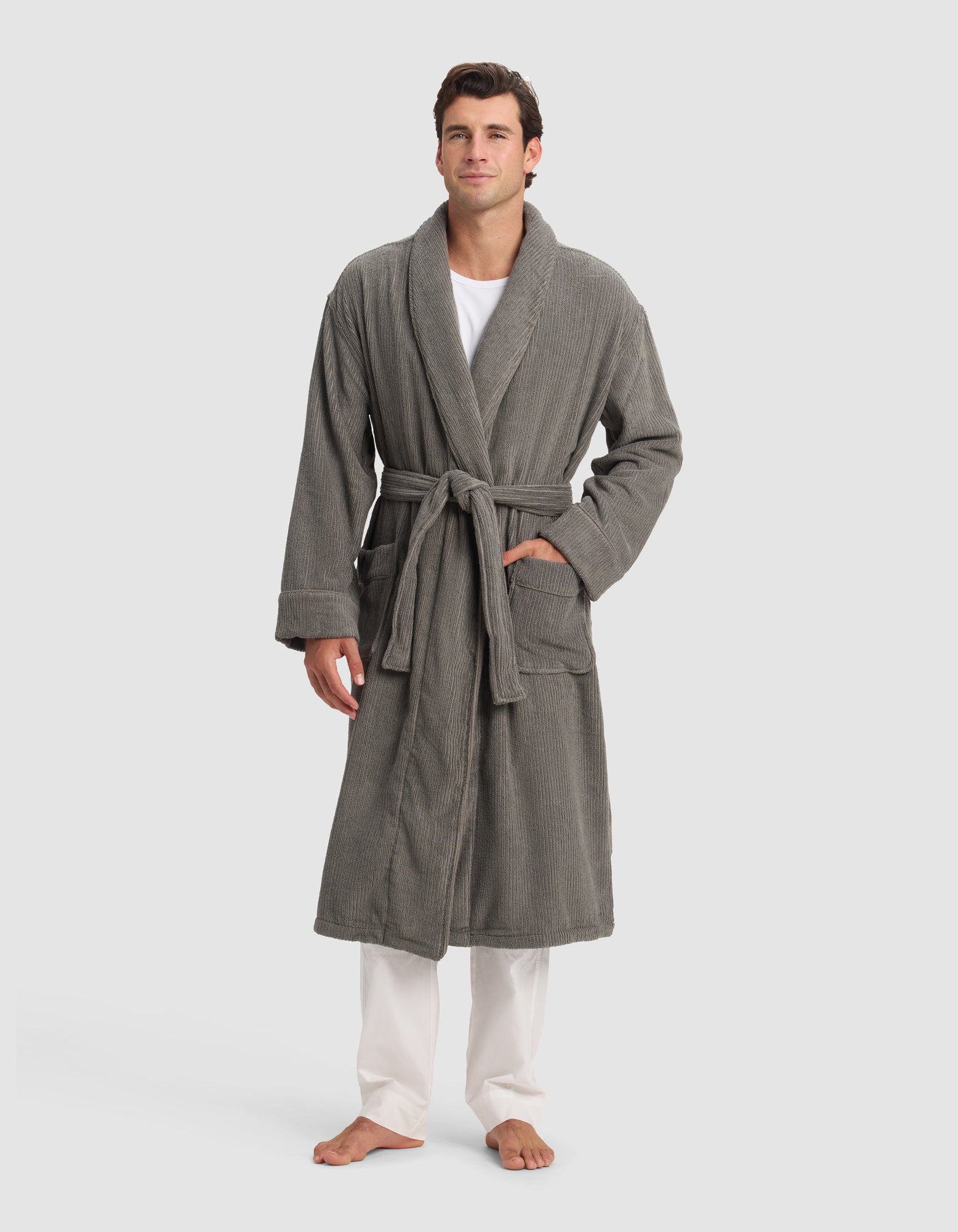 A man stands barefoot against a plain background wearing the Cozy Earth Ribbed Terry Bath Robe in gray, featuring a belt and two front pockets, layered over a white shirt and white pants.
