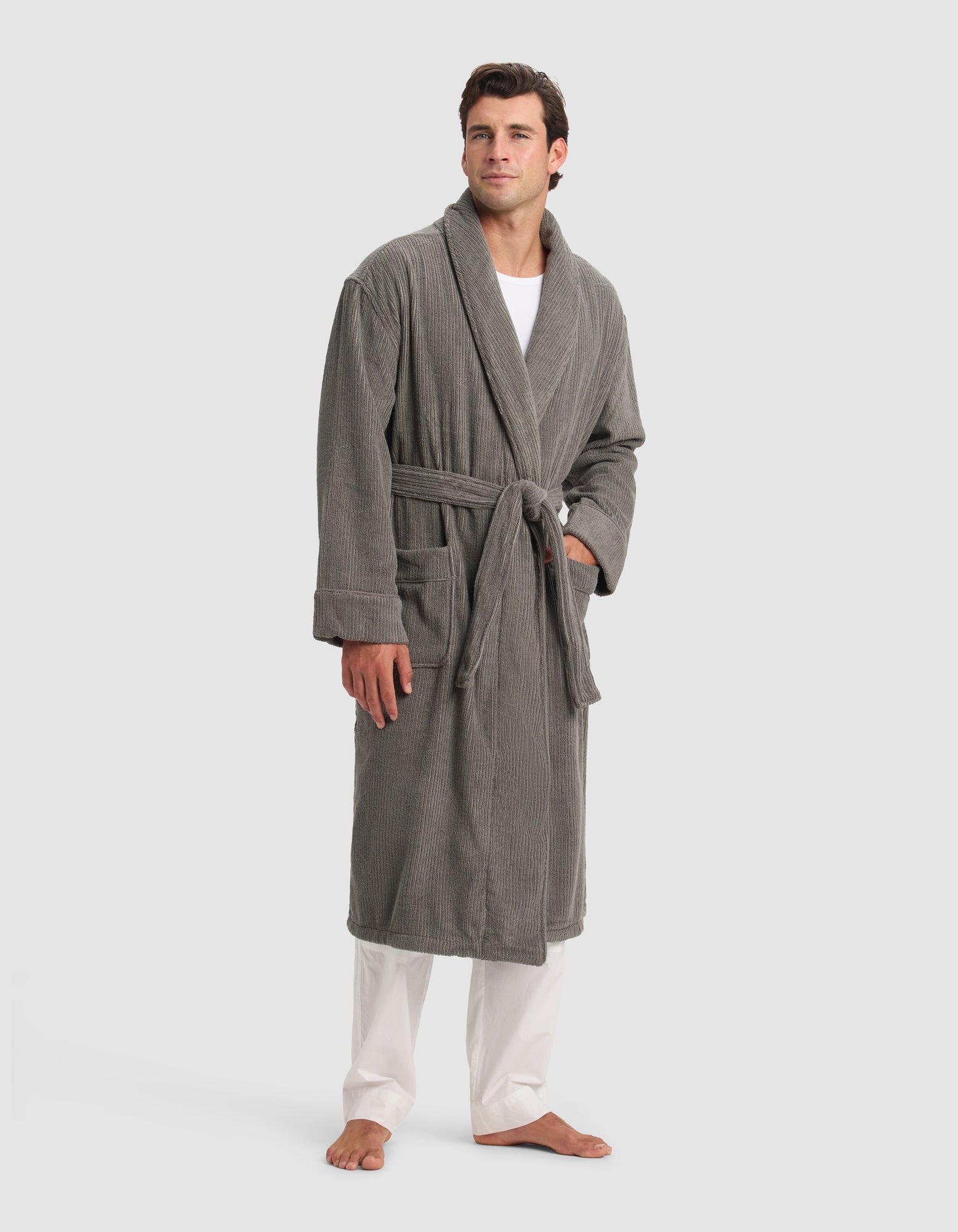 A man stands barefoot on a white background wearing the Cozy Earth Ribbed Terry Bath Robe in gray, with the belt tied at the waist. He has short brown hair and wears white pants and a white shirt underneath the robe.
