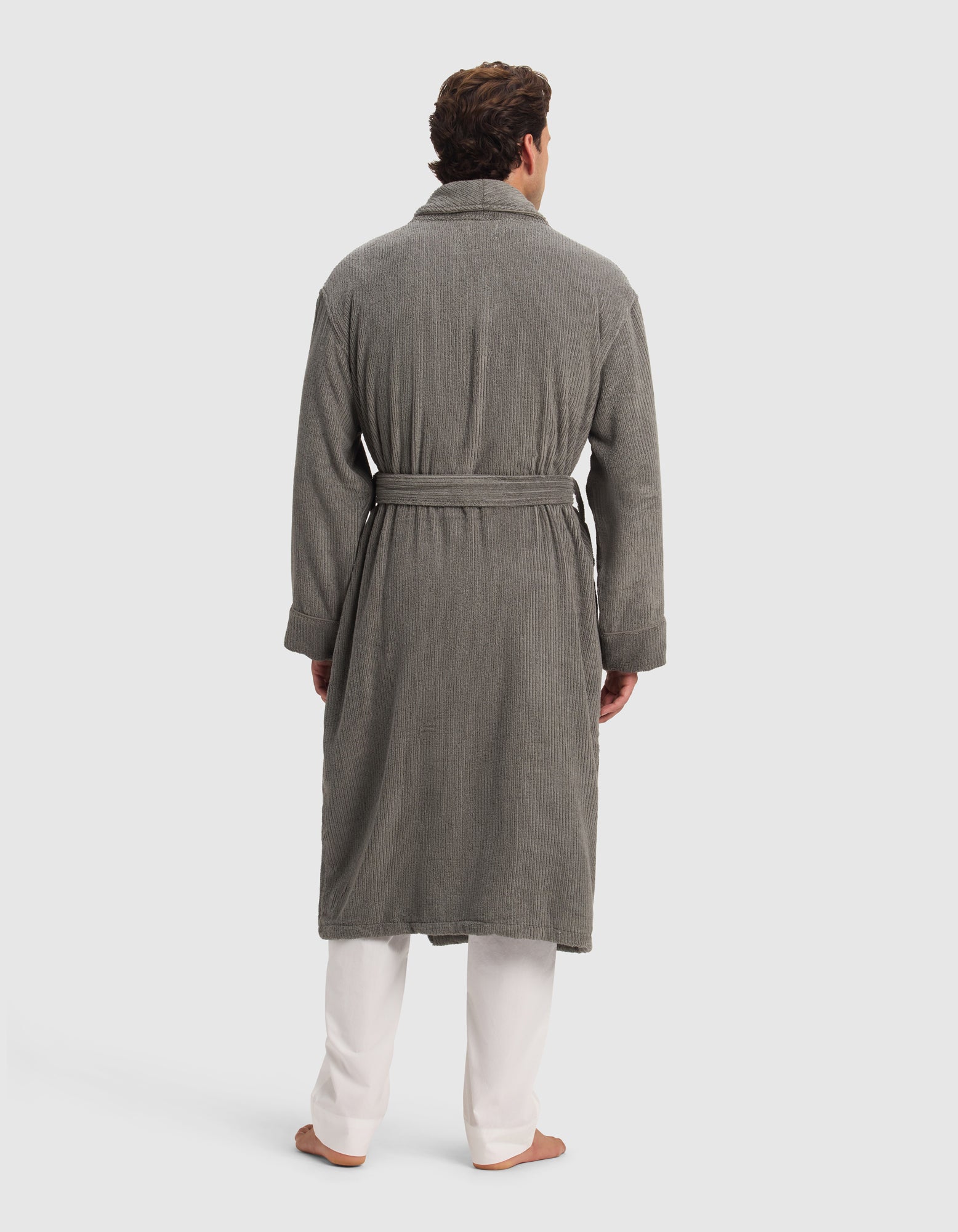 A man, barefoot against a plain light background, is wearing the Cozy Earth Ribbed Terry Bath Robe in gray, featuring a textured finish and waist belt, styled over white pants.