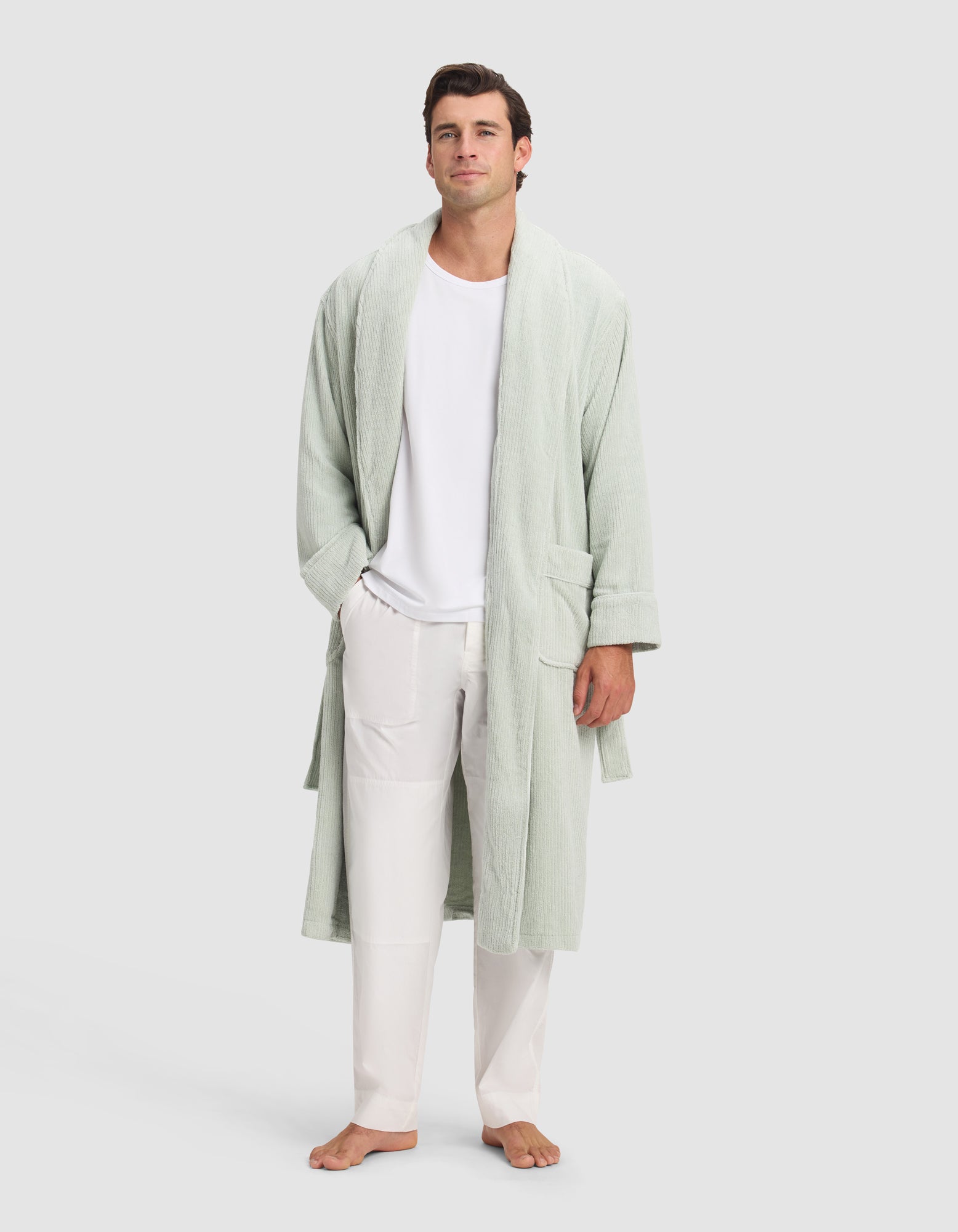 A man stands barefoot against a light background, wearing the Cozy Earth Ribbed Terry Bath Robe in light green over a white T-shirt and white pants, with his hands in the robe's pockets.