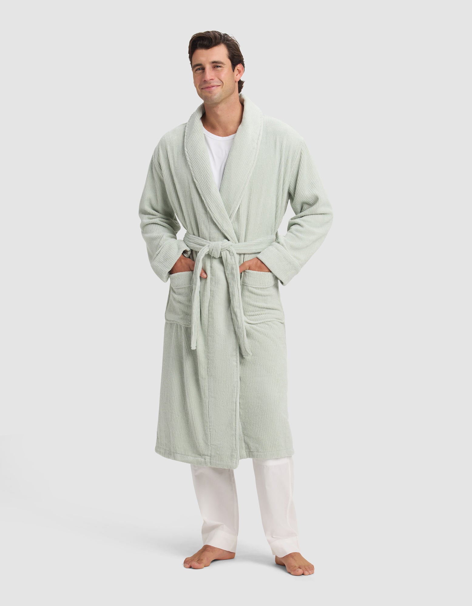 A man smiles barefoot in a Cozy Earth Ribbed Terry Bath Robe—light green with a belt and two pockets—styled over a white shirt and pants, set against a plain light background.