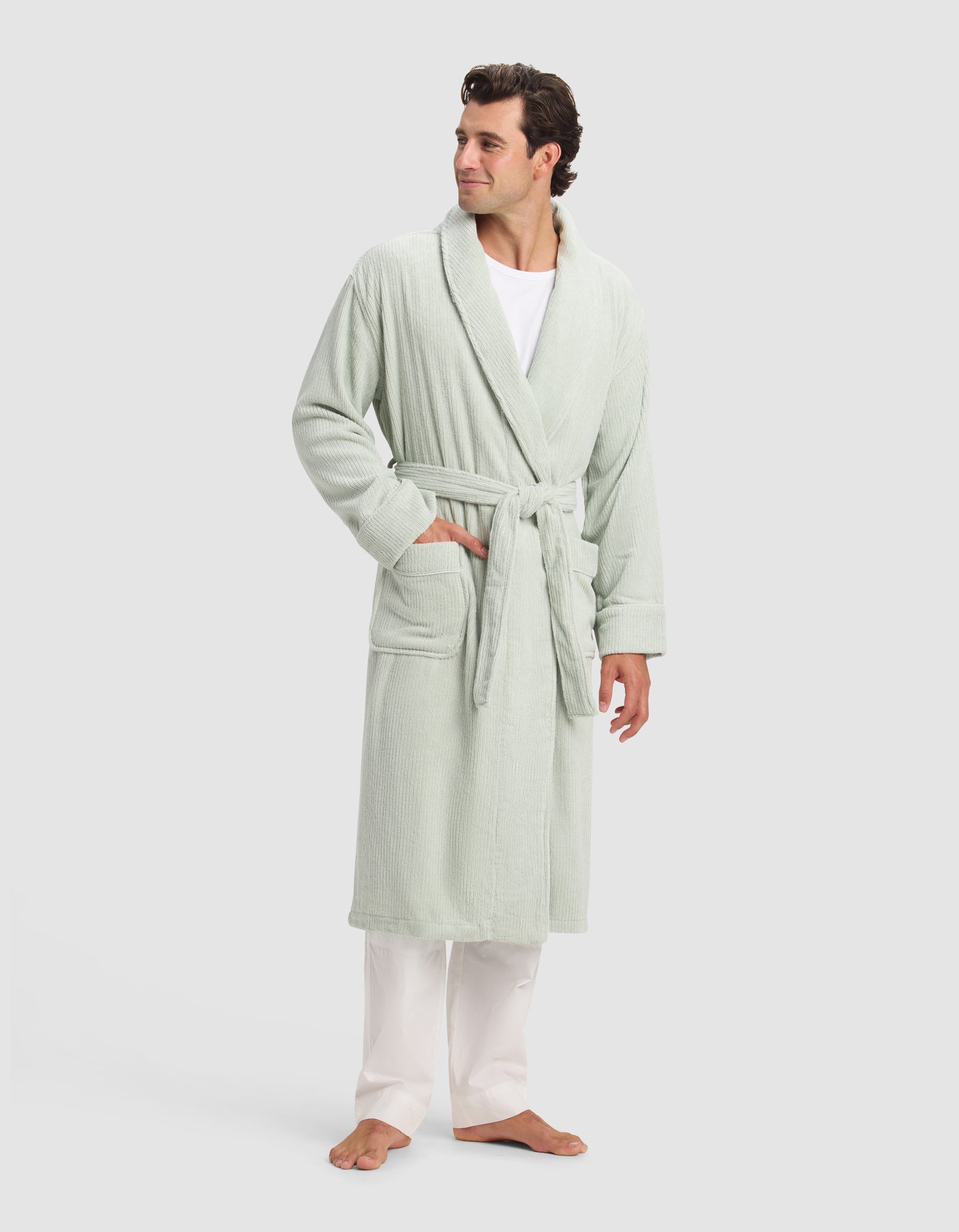 A man stands barefoot, wearing Cozy Earth's Ribbed Terry Bath Robe in light green, belted over a white shirt and pants. One hand is in his pocket as he looks to the side against a plain, light background.
