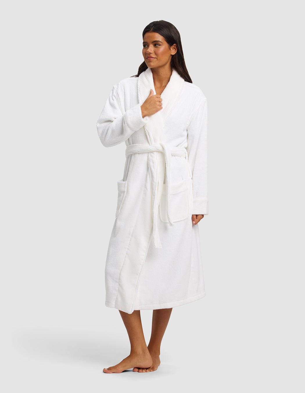 A woman stands barefoot against a light gray backdrop, wearing the Cozy Earth Ribbed Terry Bath Robe with long sleeves, large front pockets, and a belt. She looks to the side while holding the robe closed with one hand.|Color:White
