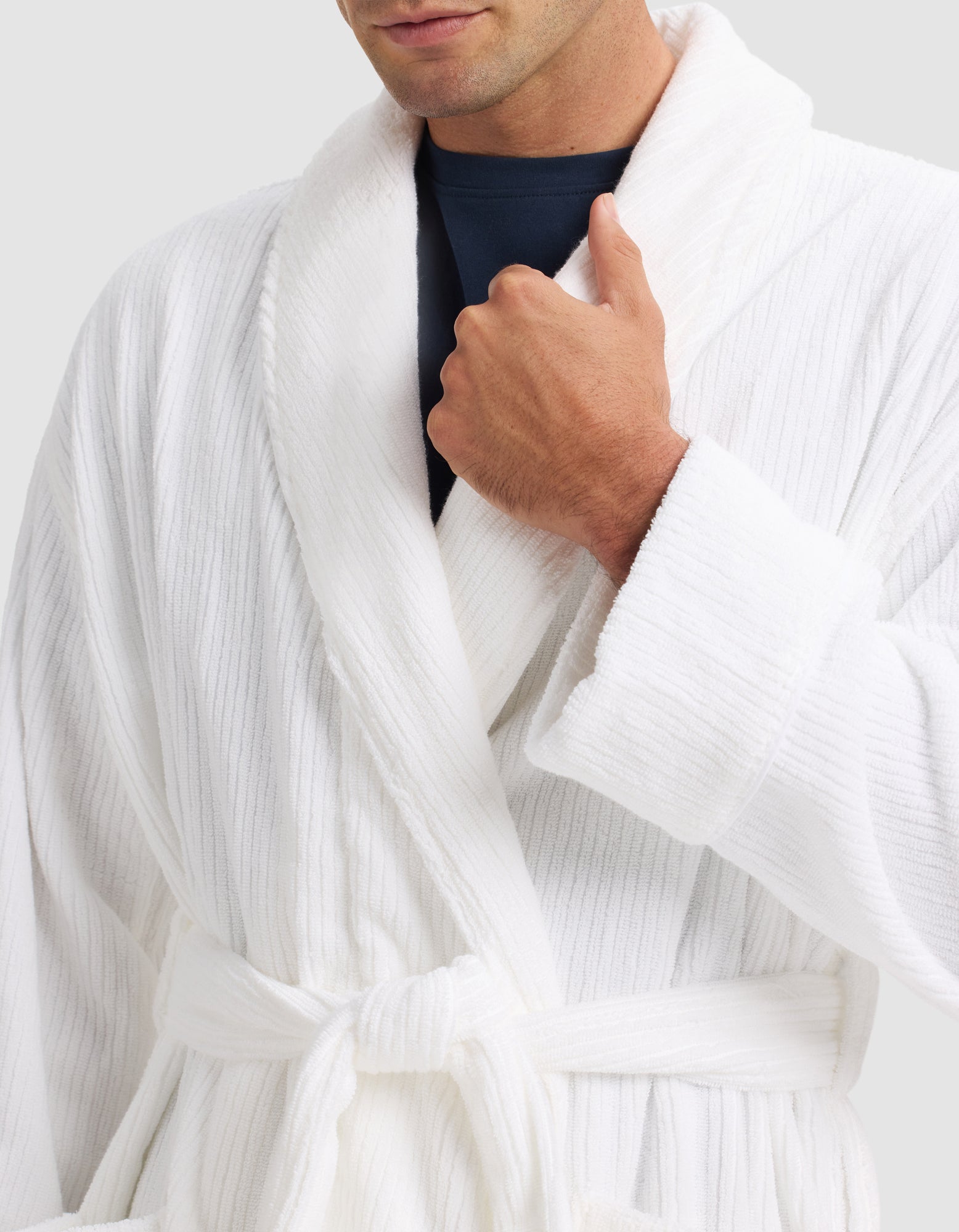 Someone wearing the Cozy Earth Ribbed Terry Bath Robe in white over a dark shirt, tying the belt at the waist and holding the collar with one hand. The person's face is partly visible.