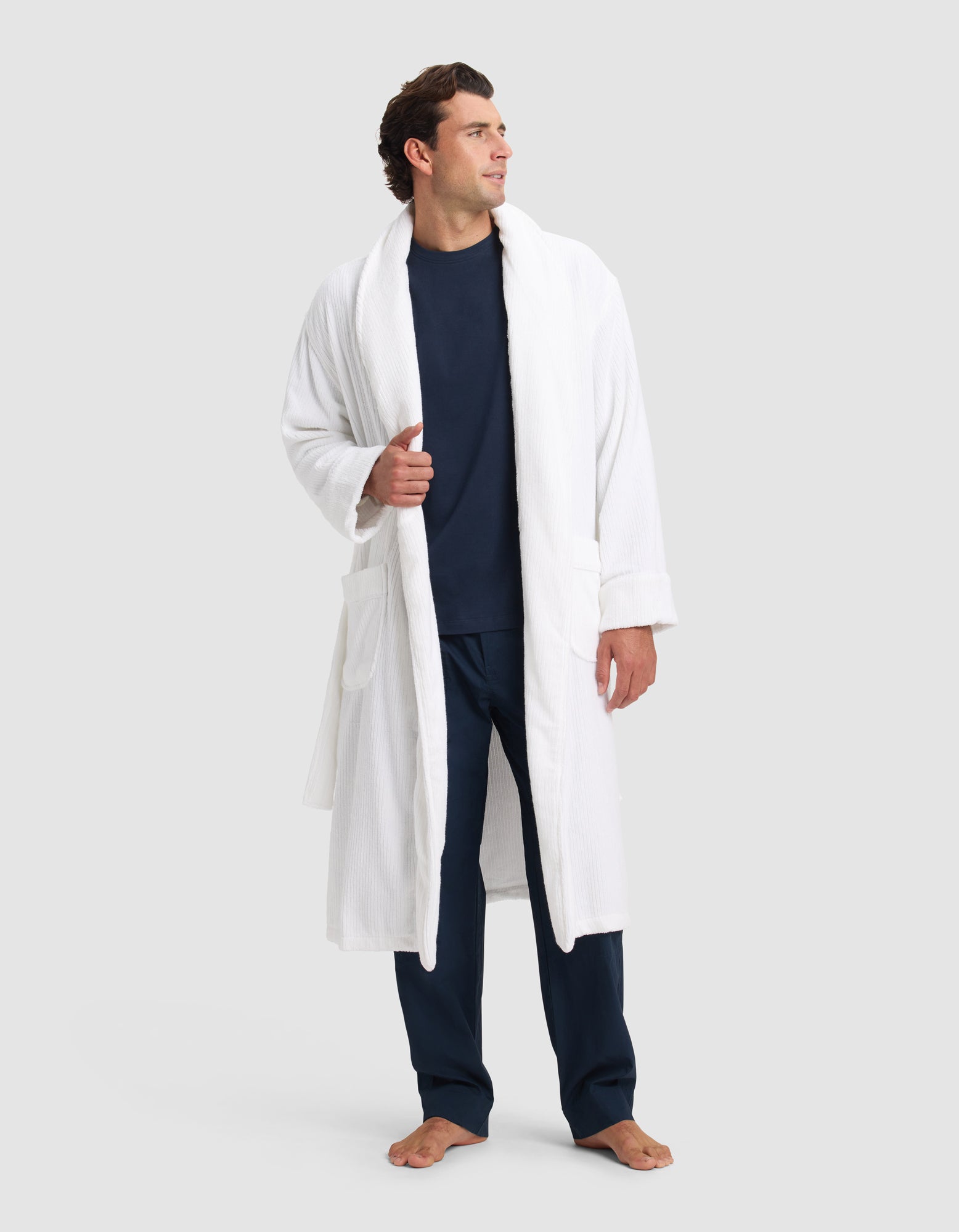 A man stands barefoot in a Cozy Earth Ribbed Terry Bath Robe over a navy shirt and pants, looking left with one hand holding the robe open. The background is plain light gray.