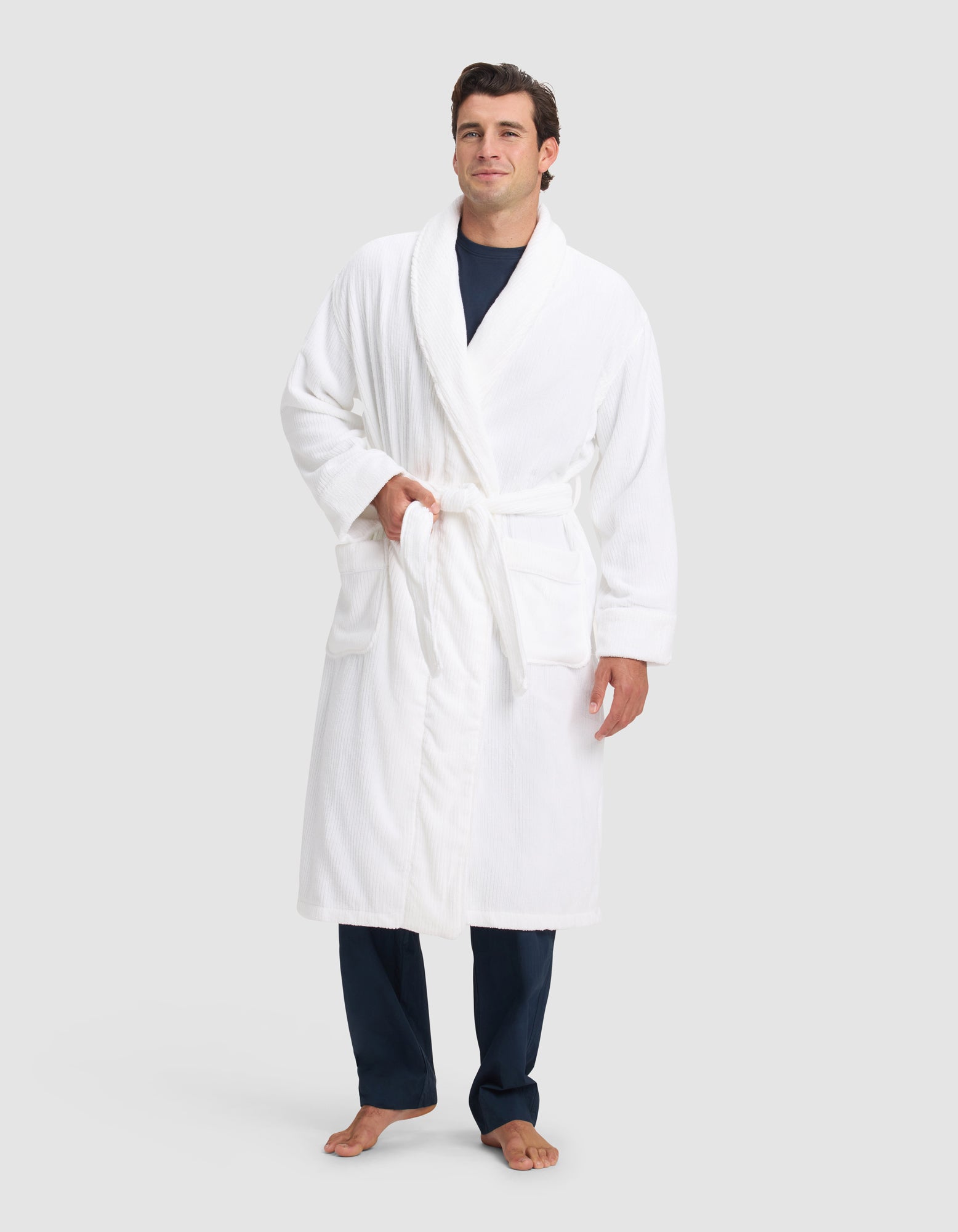 A person stands barefoot in a Cozy Earth Ribbed Terry Bath Robe over dark blue pajamas, posing against a light background with one hand in a pocket and the other at their side.