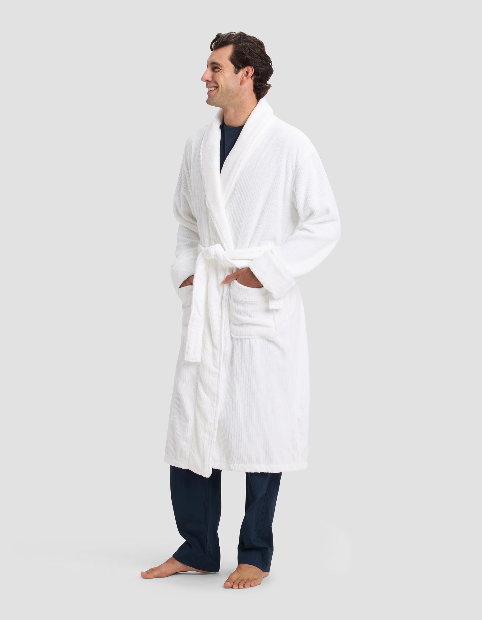 A man stands barefoot, smiling, with his hands in the pockets of a Cozy Earth Ribbed Terry Bath Robe tied at the waist over navy pants and a navy shirt. The background is plain light gray.