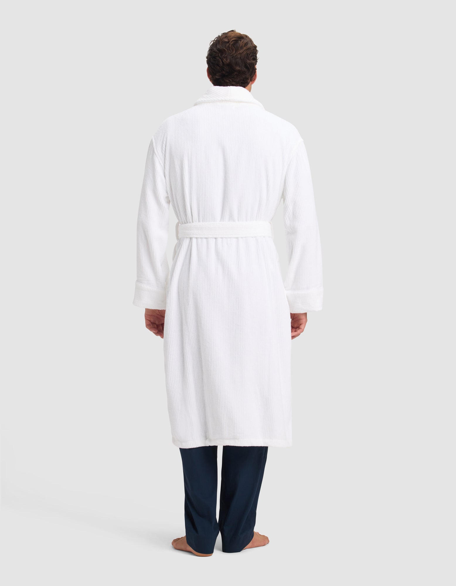 A person faces away, wearing the Cozy Earth Ribbed Terry Bath Robe with a belt, dark pants, and bare feet, set against a plain light background.