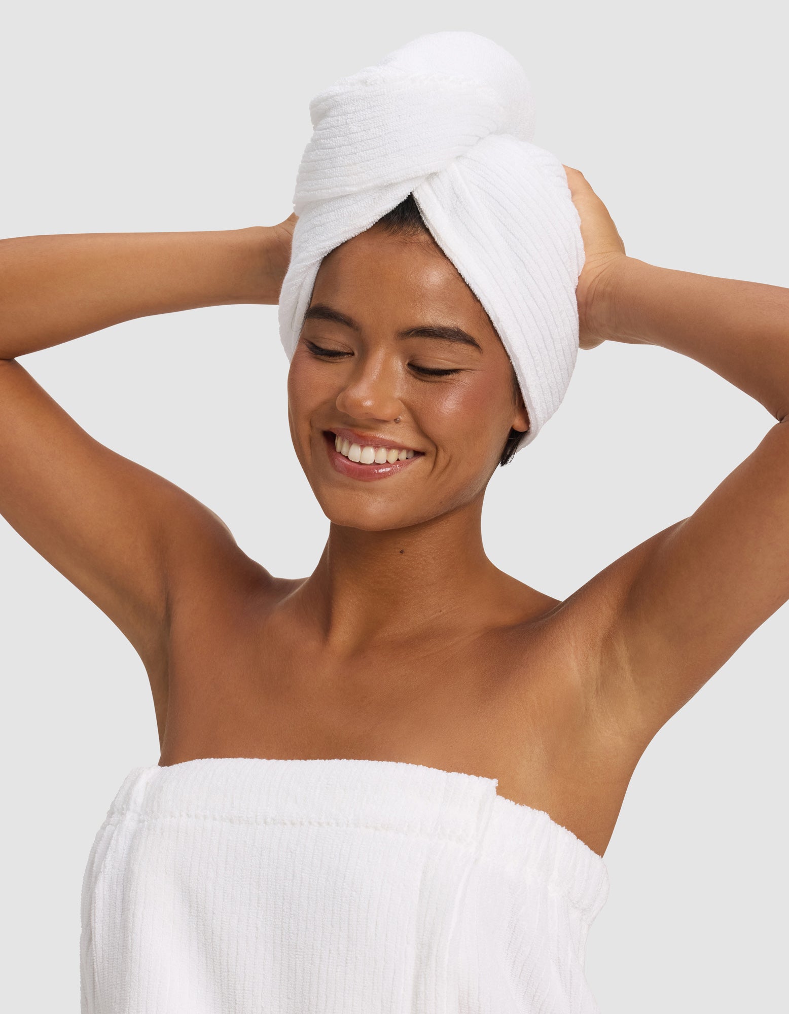 A smiling woman poses with arms raised, wearing a Cozy Earth Ribbed Terry Hair Towel and a white towel wrapped around her body, standing against a plain light gray background.|Color:White