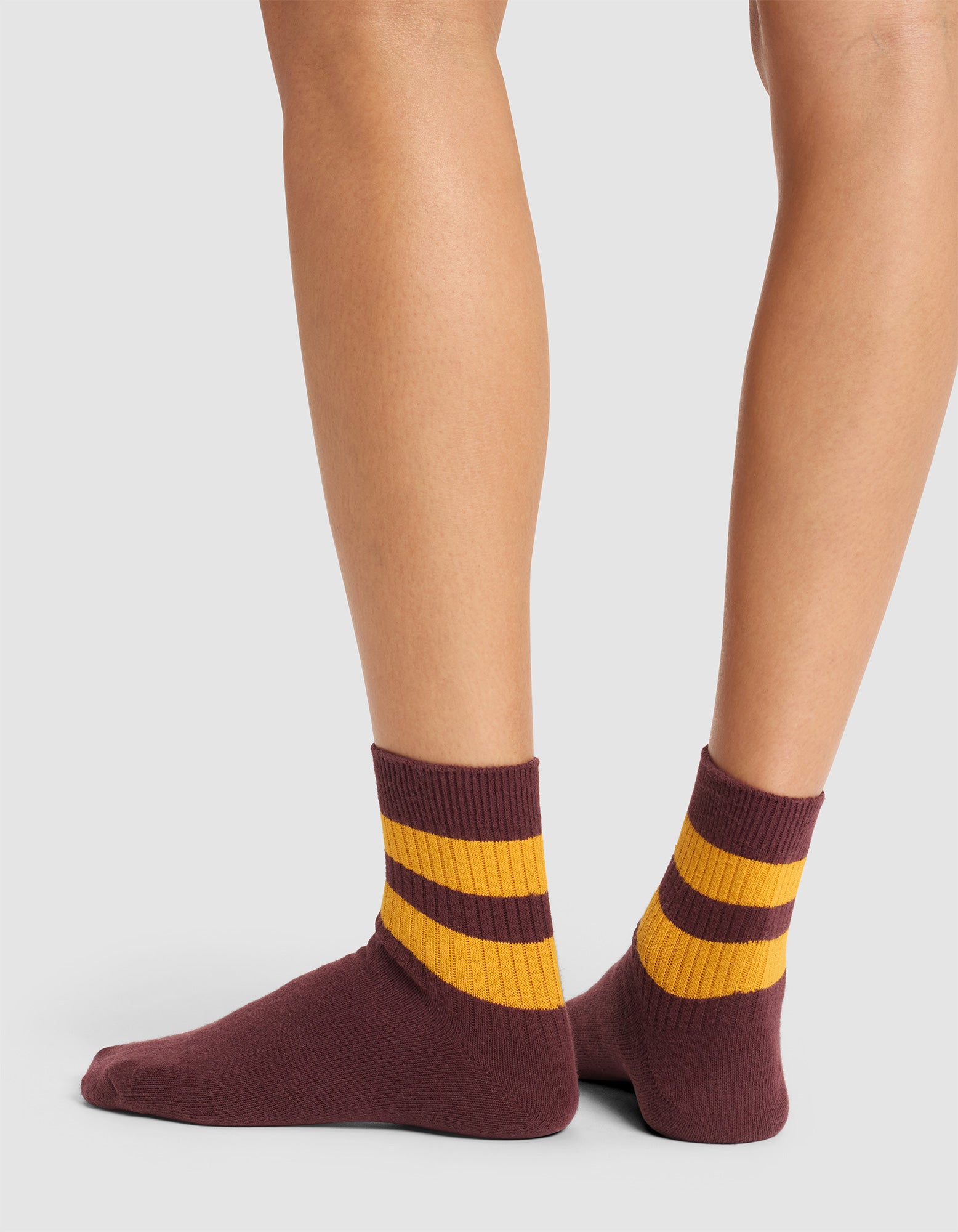A person wearing Cozy Earth Women's Sadie Socks, featuring maroon with two thick yellow stripes, stands on a light gray surface seen from behind at ankle level. |Color:Burgundy/Goldenrod