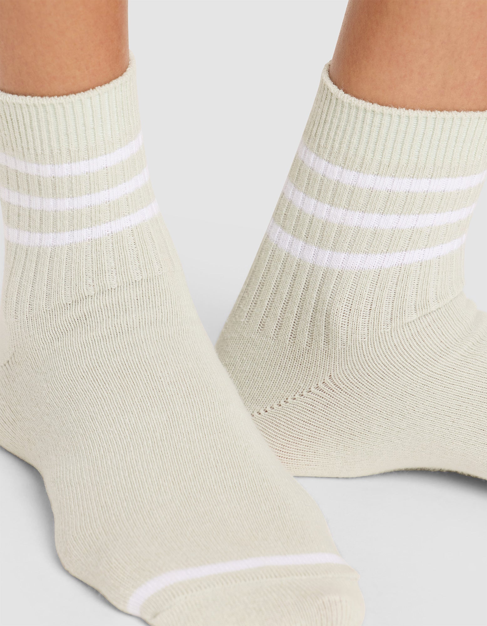 Close-up of two feet wearing Cozy Earth Women's Sadie Socks in light beige with ribbing and three horizontal white stripes at the top, standing on a light gray surface.