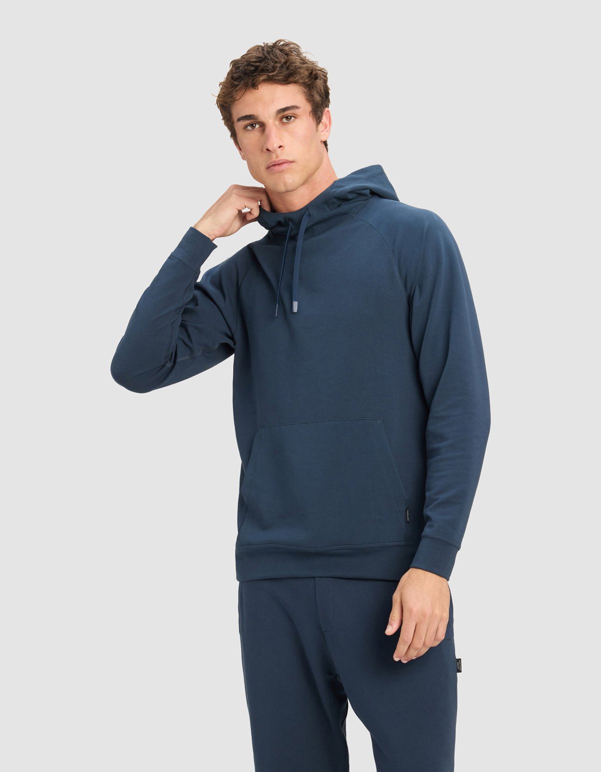 A young man models Cozy Earth's Men's StretchTech Hoodie in dark blue with matching pants, standing against a light gray background, one hand near his neck and the other by his side.|Color:Eclipse