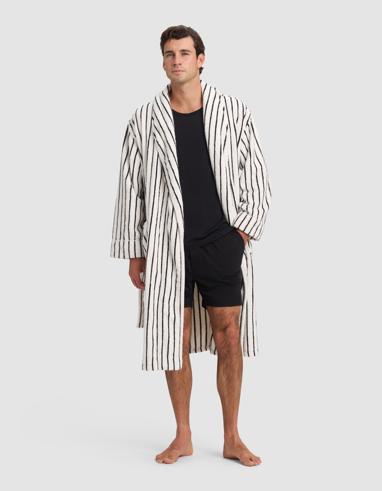 A man stands barefoot against a plain background, wearing the knee-length Luxe Bath Robe by Cozy Earth in white with black vertical stripes, layered over a black t-shirt and shorts. One hand is in his pocket and he looks neutral.