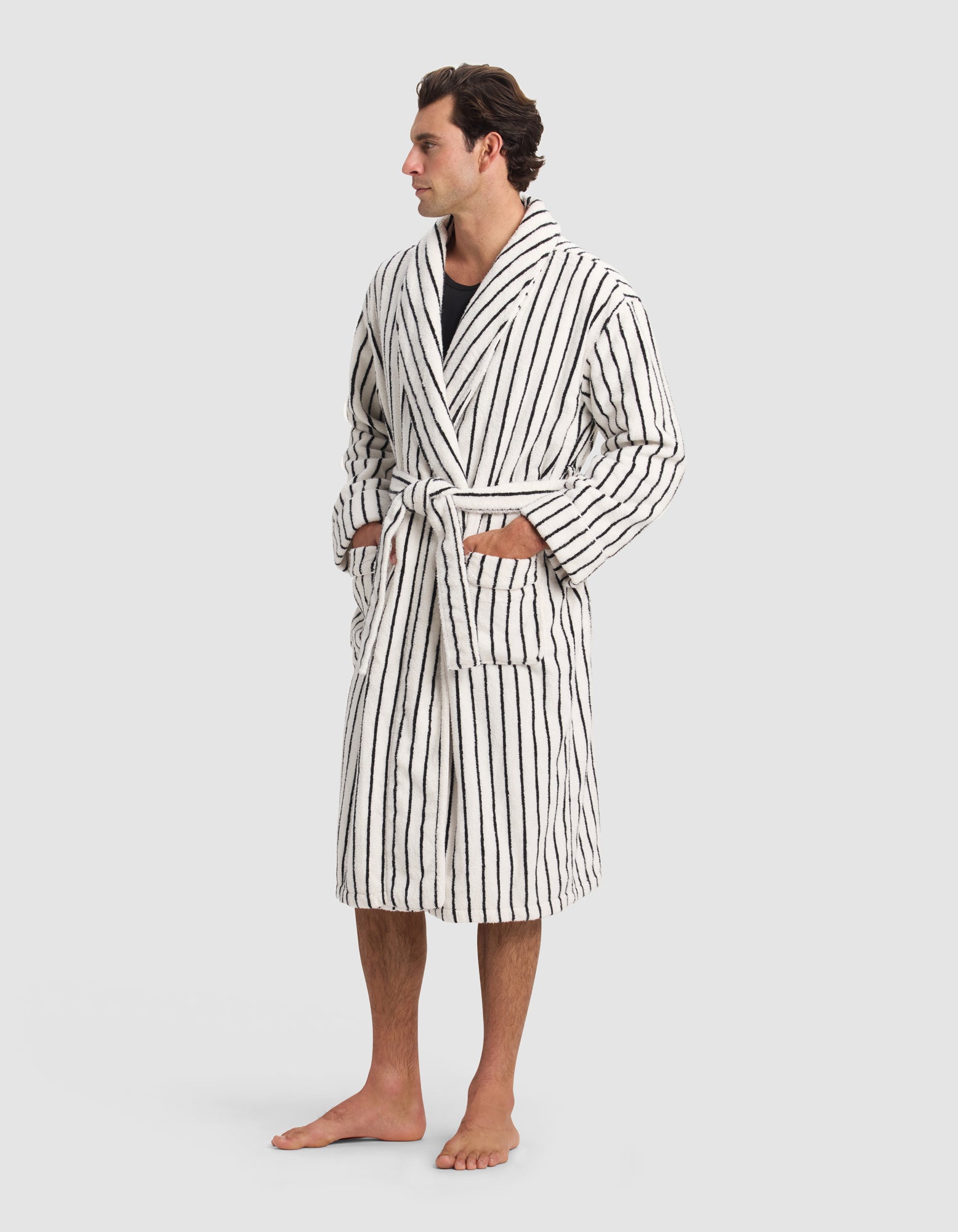 A man stands barefoot in the Cozy Earth Luxe Bath Robe—white with vertical black stripes and a matching belt—hands in front pockets, looking to the side against a plain light gray background.