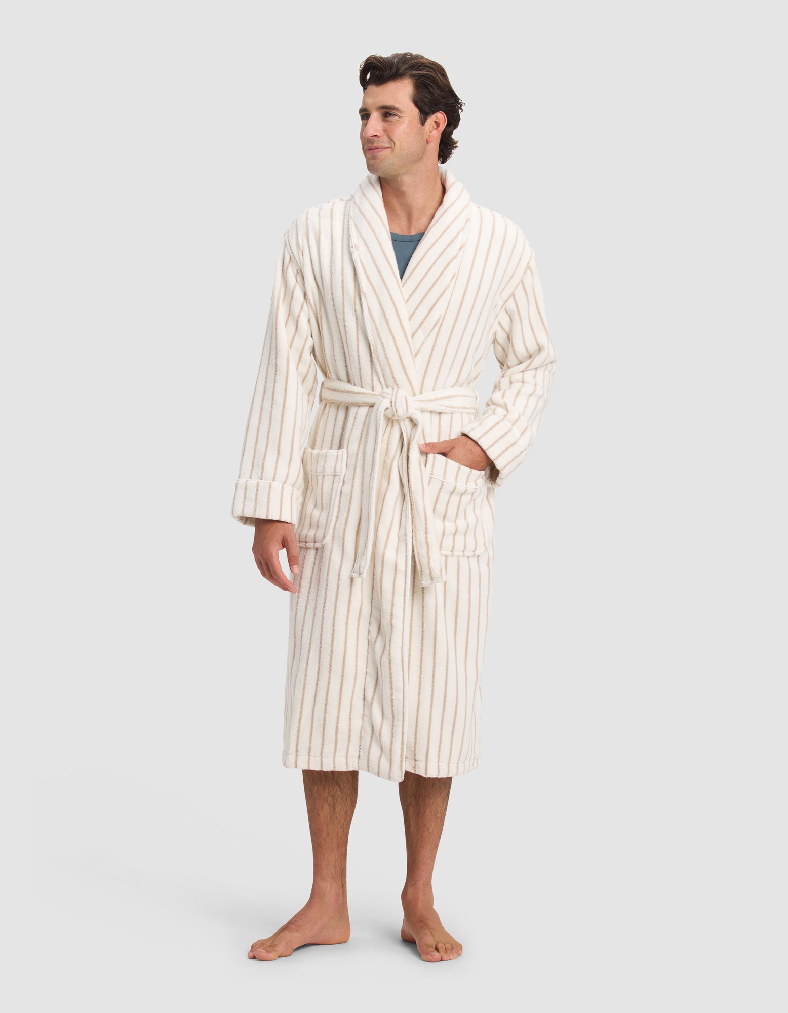 A man stands barefoot, smiling slightly and looking to the side, wearing Cozy Earth's Luxe Bath Robe in cream with thin beige stripes, a belted waist, two front pockets, and a gray shirt underneath.