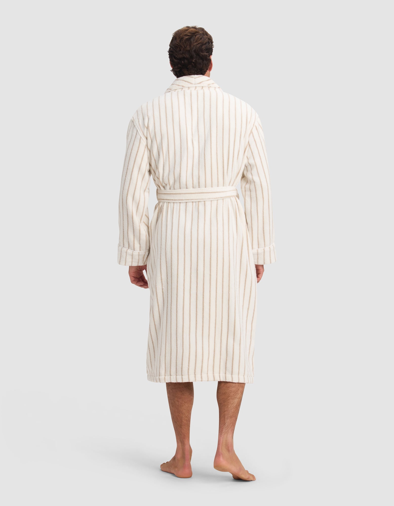 A man stands barefoot facing away, wearing the Cozy Earth Luxe Bath Robe—white with thin beige stripes and a belted waist—against a plain light gray background.