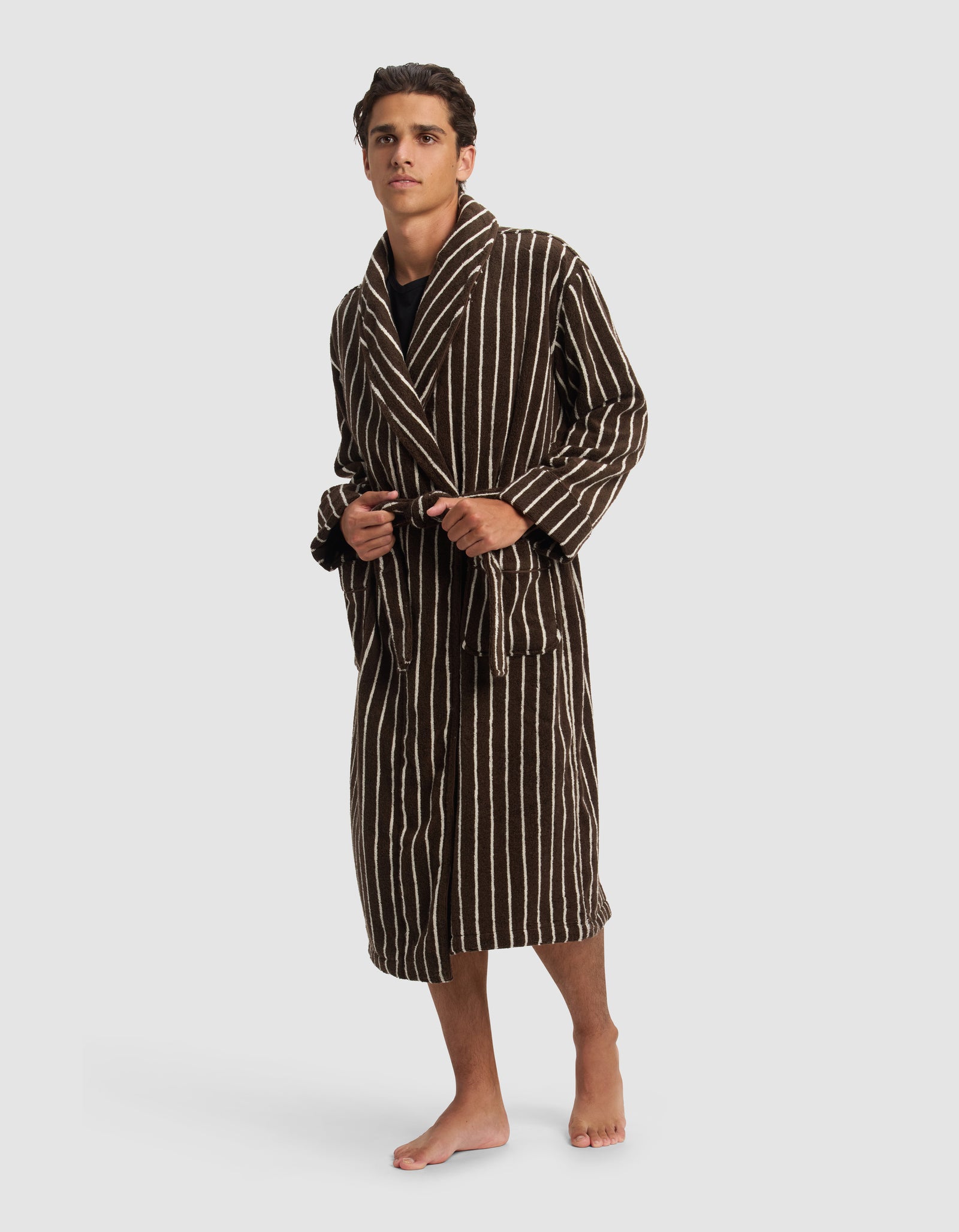 A young man stands barefoot, slightly turned to the side, modeling the Cozy Earth Striped Luxe Bath Robe—a knee-length brown robe with white vertical stripes and a waist belt—against a plain light background.