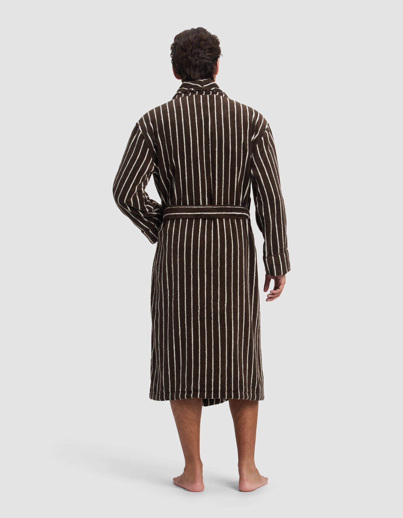 A person stands barefoot, facing away, wearing Cozy Earth's Striped Luxe Bath Robe featuring vertical white stripes and a waist tie.