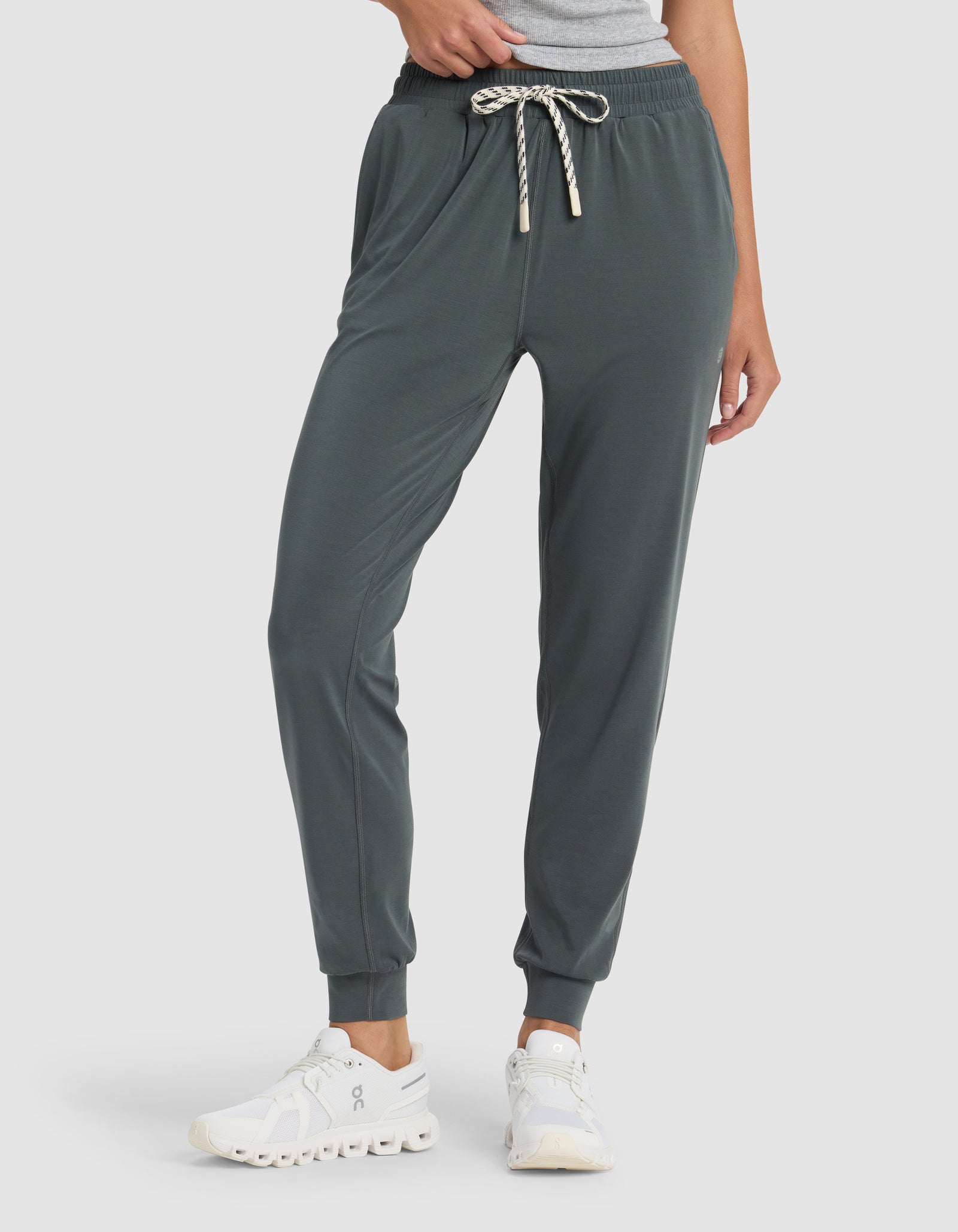 A person wears Cozy Earth's Women's Studio Jogger in dark green with a striped drawstring, white sneakers, and a gray top. The photo captures the outfit from the waist down against a plain background.