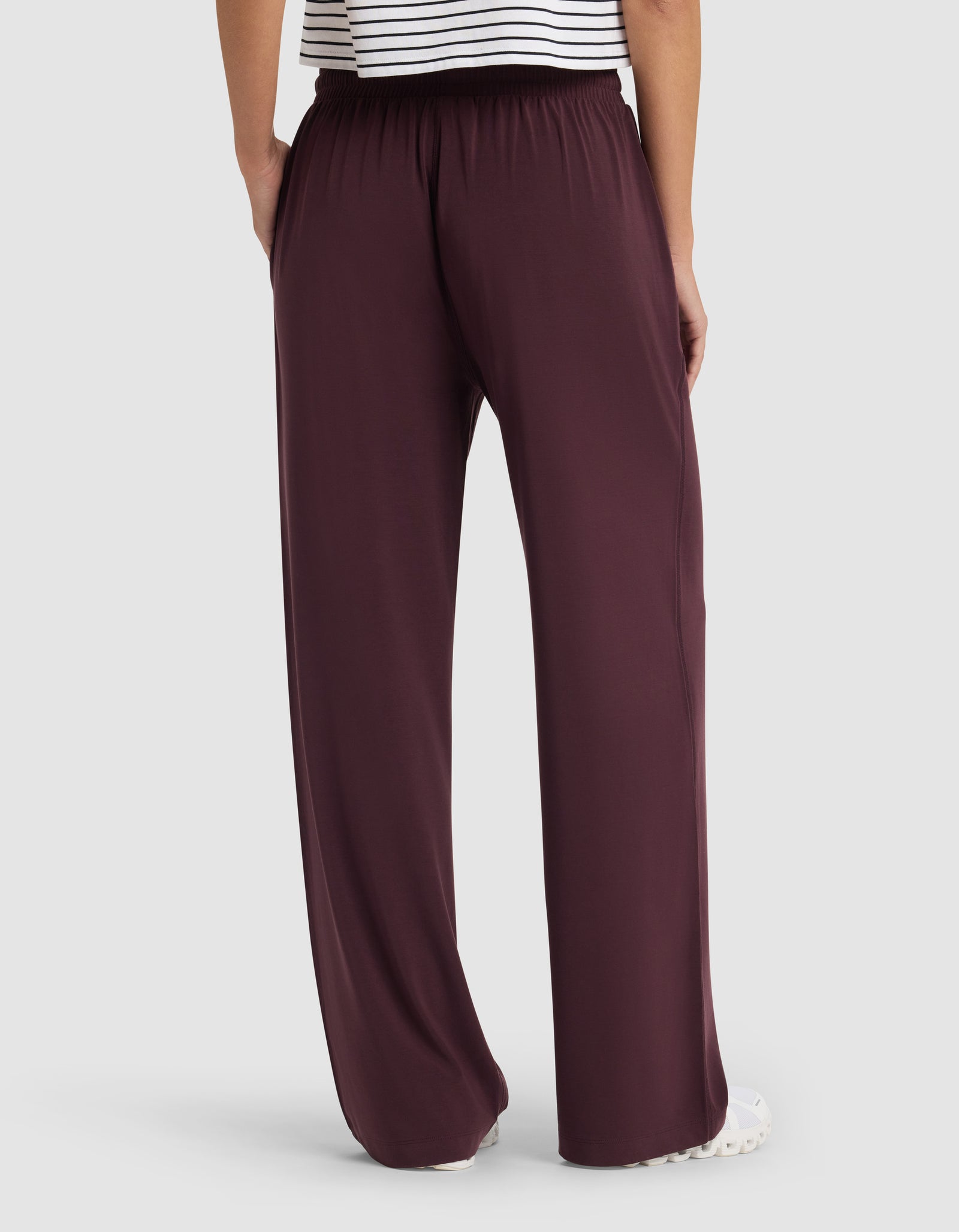 A person faces away, showing only their lower back and legs, wearing Cozy Earth's Women's Studio Wide Leg Pant in dark burgundy with an elastic waistband, paired with a white top featuring black stripes.