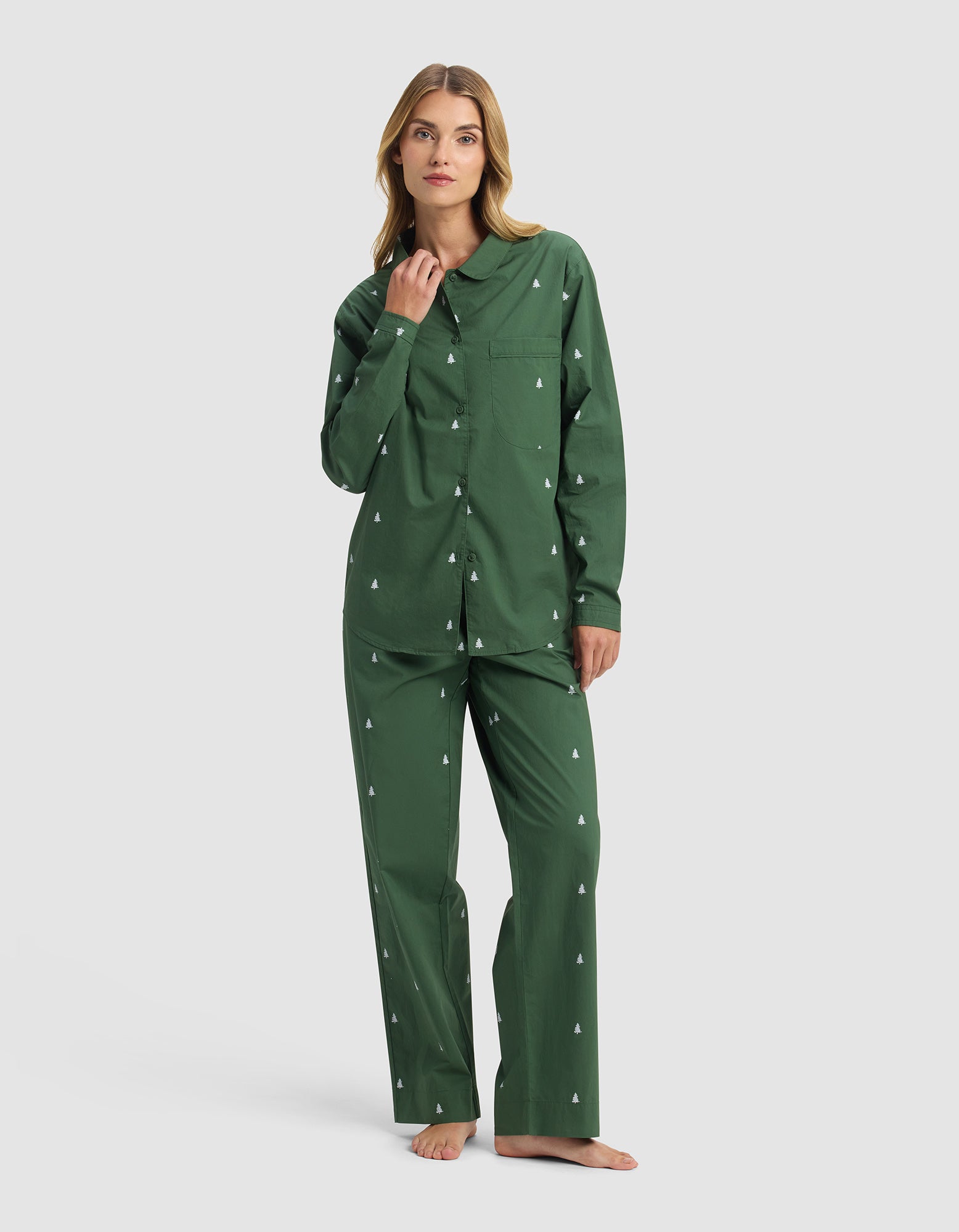 A woman stands barefoot in Cozy Earth's Women's Sutton Long Sleeve Pajama Set, featuring green pajamas with a small white tree pattern, a button-up long-sleeve shirt, and matching pants, against a plain white background.|Color:Jade Tree