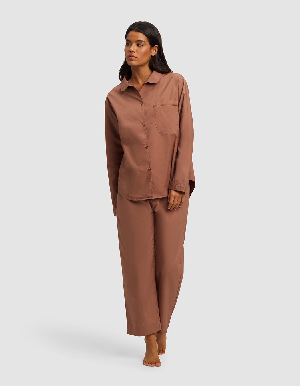 A woman stands barefoot against a plain background, wearing the Cozy Earth Women's Sutton Long Sleeve Pajama Set—a loose-fitting brown button-up top with a chest pocket and matching wide-leg pants.|Color:Saddle