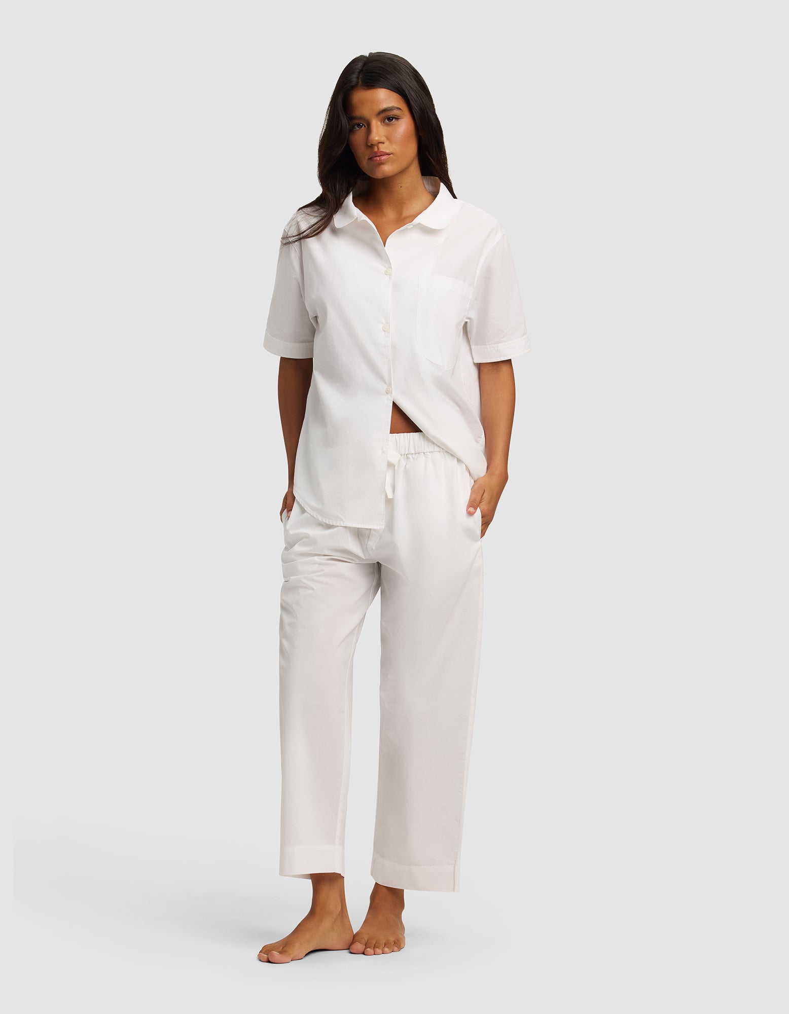 A woman stands barefoot in the Cozy Earth Women's Sutton Short Sleeve & Pant Pajama Set, featuring a matching white button-up shirt and relaxed-fit pants. She has long dark hair, hands in her pockets, against a plain light gray background.|Color:White