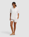 A woman stands barefoot against a plain background, wearing the Cozy Earth Women's Sutton Short Sleeve Pajama Set—featuring a loose-fitting white button-up shirt and matching shorts—with one hand in her pocket and a relaxed expression.|Color:White