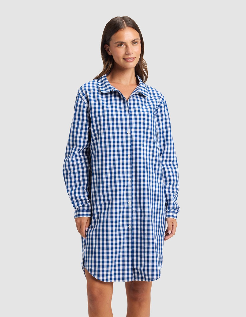 A woman with straight brown hair wears the Cozy Earth Women's Sutton Sleep Dress, a blue and white checkered, long-sleeve shirt dress that falls above the knees. She smiles slightly against a plain light background.|Color:Pacific Gingham