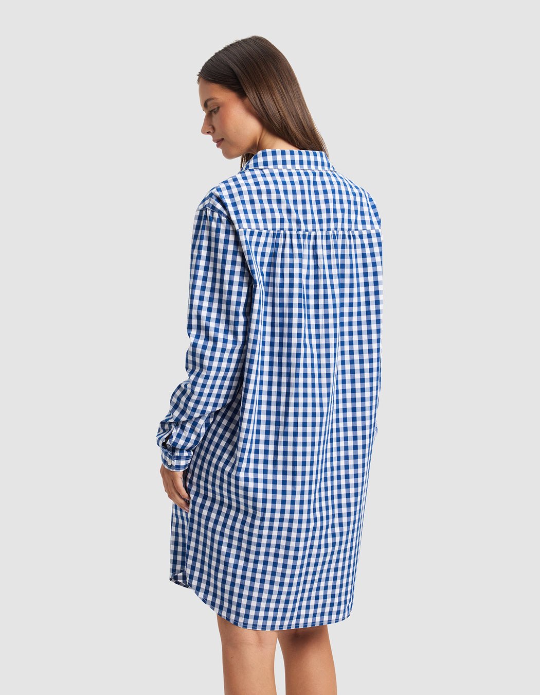A woman faces away wearing Cozy Earth's Women's Sutton Sleep Dress—a loose-fitting, blue and white checkered long-sleeve shirt dress that falls above the knees. The background is plain light gray.|Color:Pacific Gingham