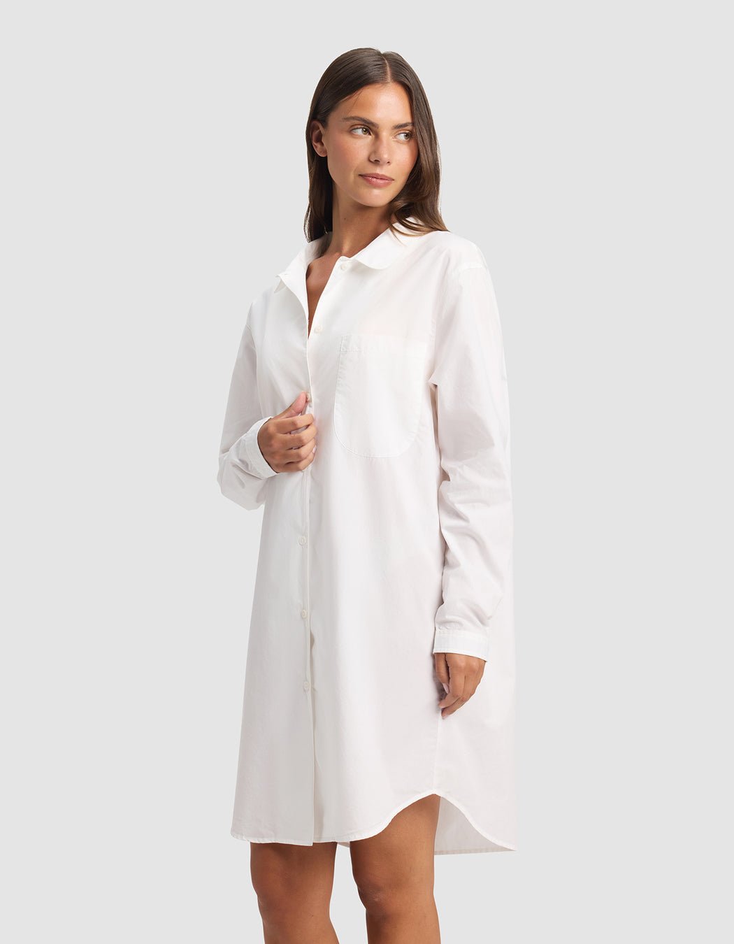 A woman in the Cozy Earth Women's Sutton Sleep Dress, a long-sleeved white button-down style, stands against a plain light gray background, gazing slightly left with a relaxed expression.|Color:White