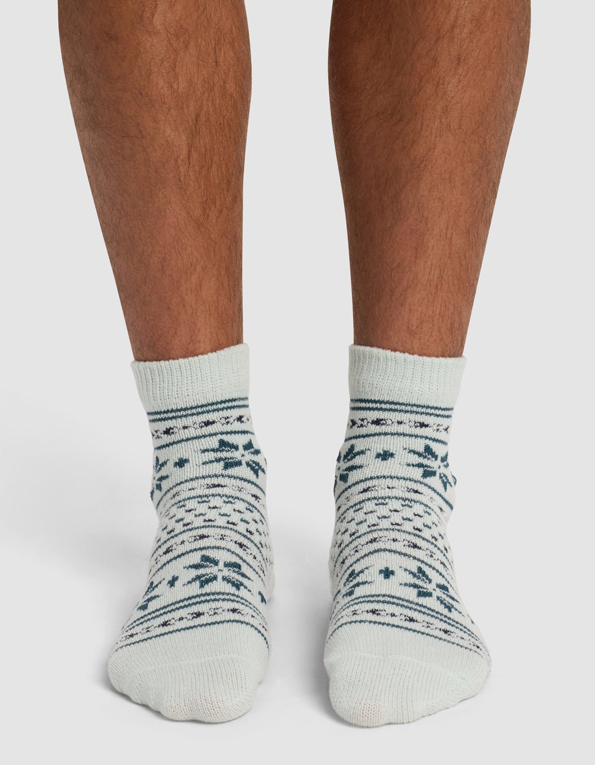 A person wearing Cozy Earth's The Snowflake Plush Modern Crew Sock—cream-colored with dark green and blue snowflake and stripe patterns—stands on a white surface.