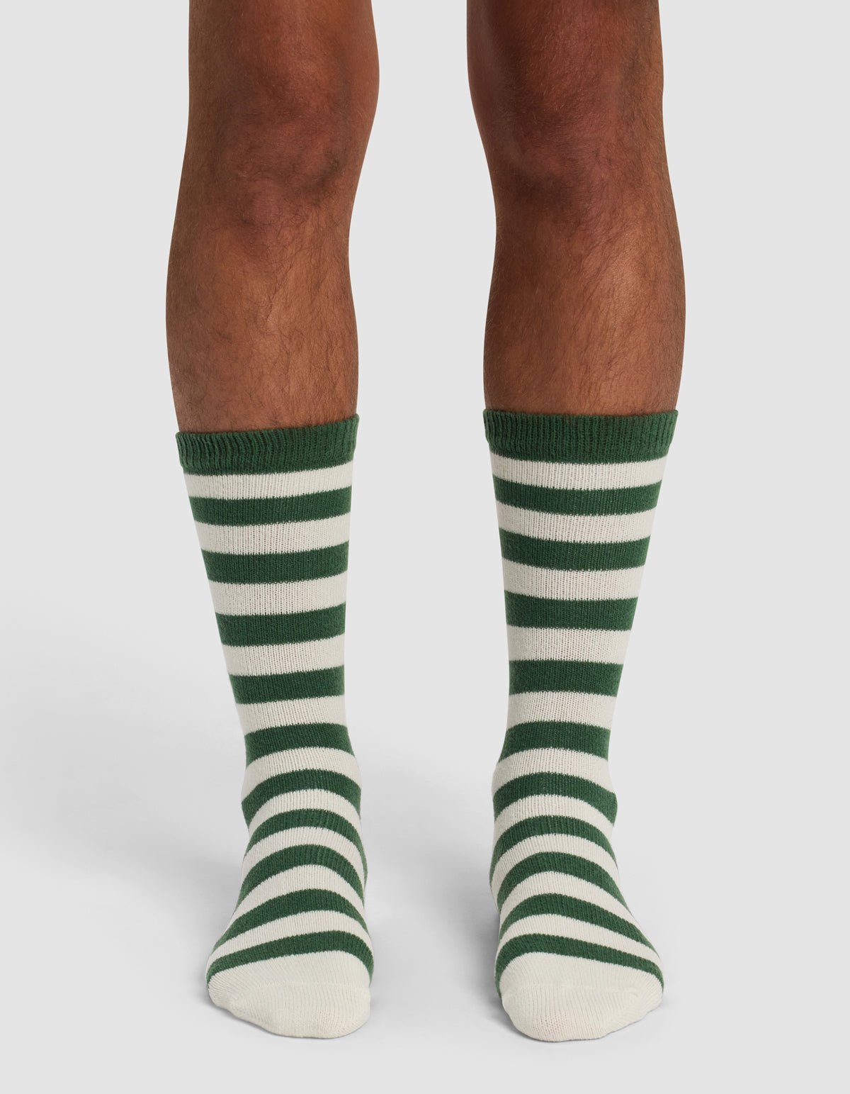 A person wearing The Striped Plush Lounge Sock by Cozy Earth, featuring knee-length horizontal green and white stripes, stands on a white background.