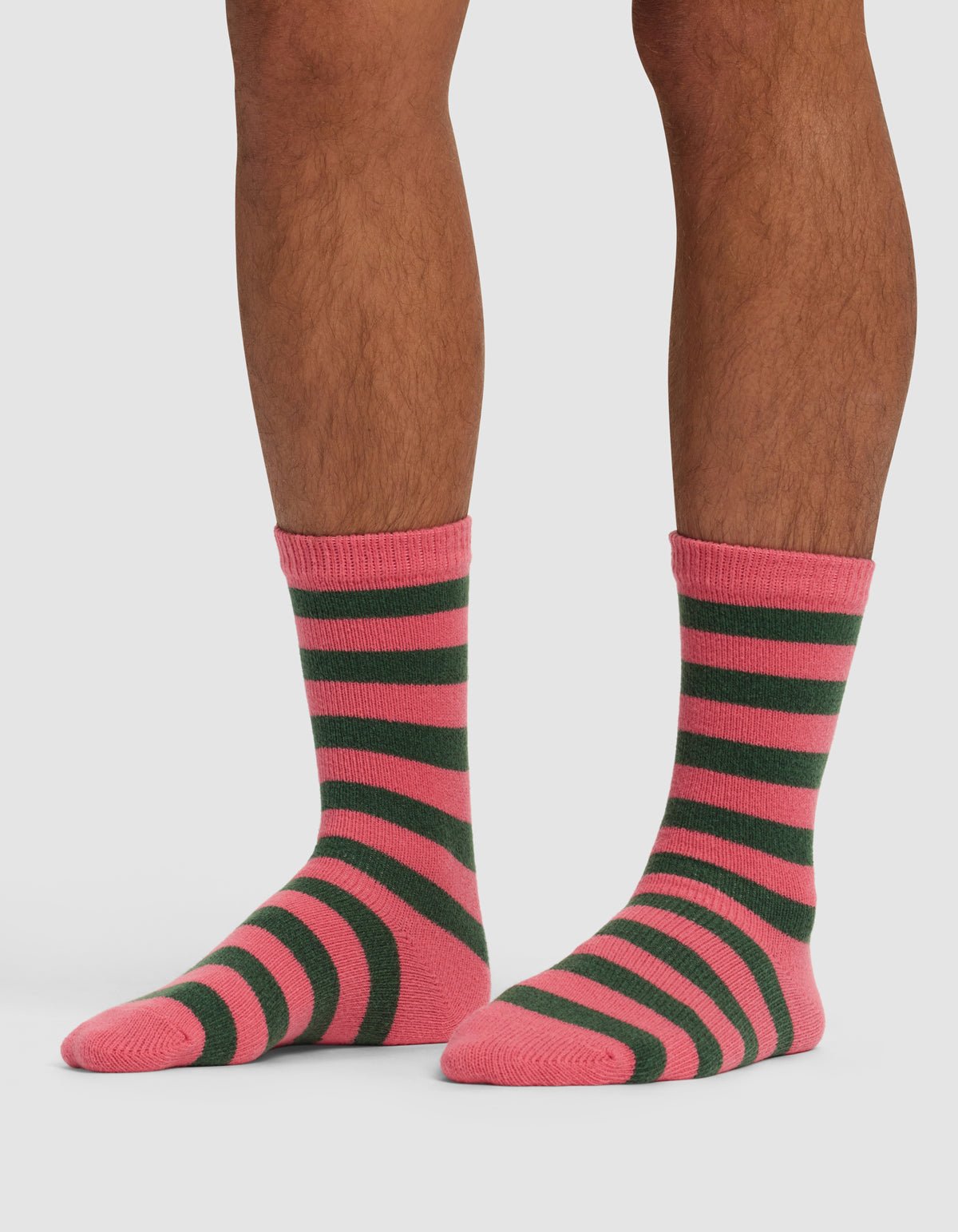 A person shows their lower legs and feet while wearing Cozy Earth's Striped Plush Lounge Sock in pink and dark green stripes, standing on a white background.