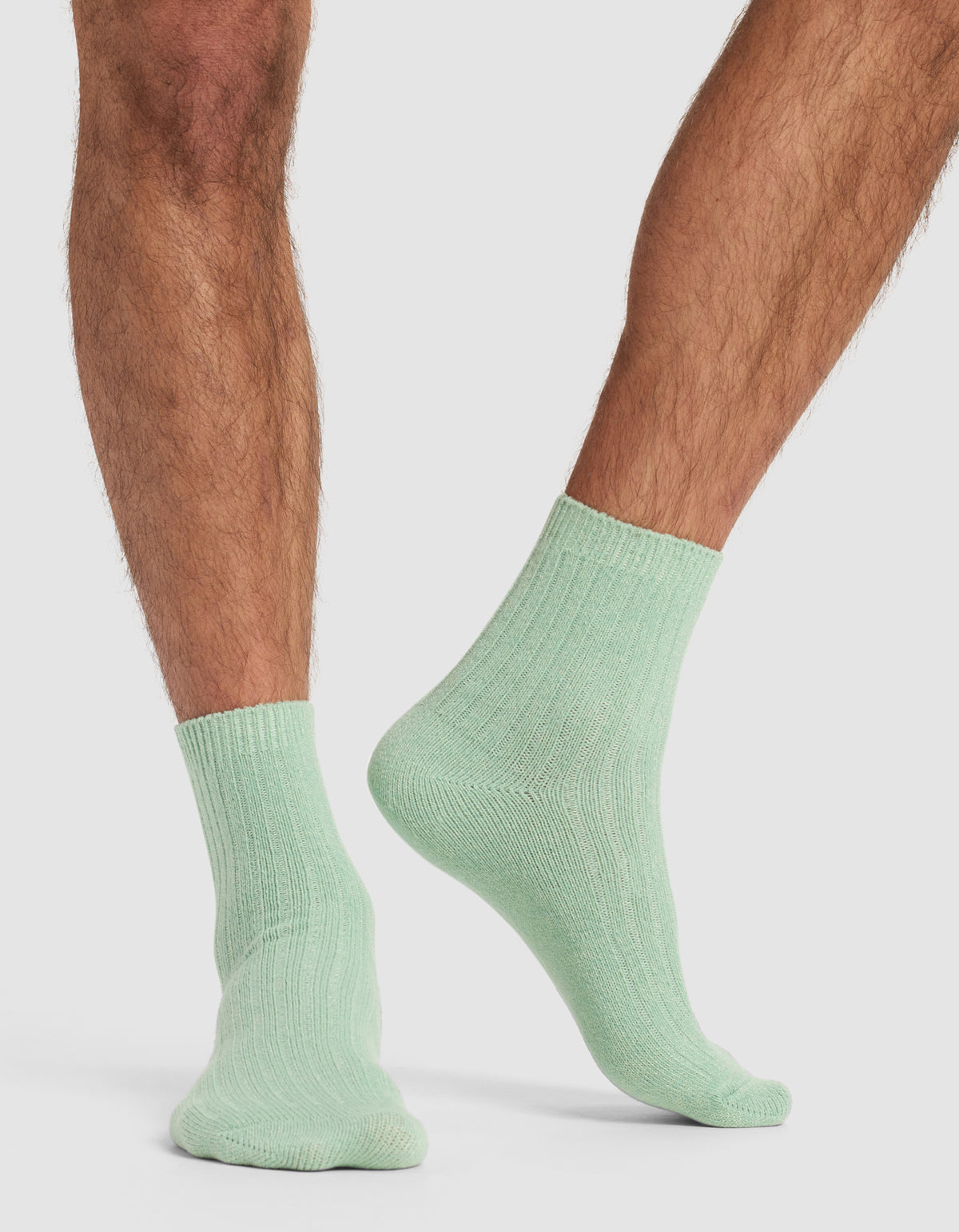 A person with light brown, hairy legs models Cozy Earth's The Plush Modern Crew Sock 3-Pack in pastel green, standing on a white background with one foot flat and the other slightly raised at the heel.