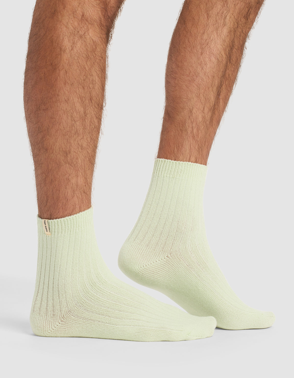 A person with hairy legs stands on a white surface wearing light green socks from Cozy Earth's The Plush Modern Crew Sock 3-Pack.